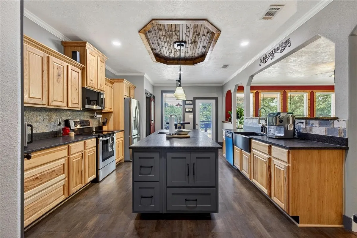 Spacious kitchen featuring a central island with integrated storage, wood-finish flooring, recessed lighting, and a wood-paneled ceiling accent. Granite counters, hickory cabinets, tile back splash, double black metal farm sink.