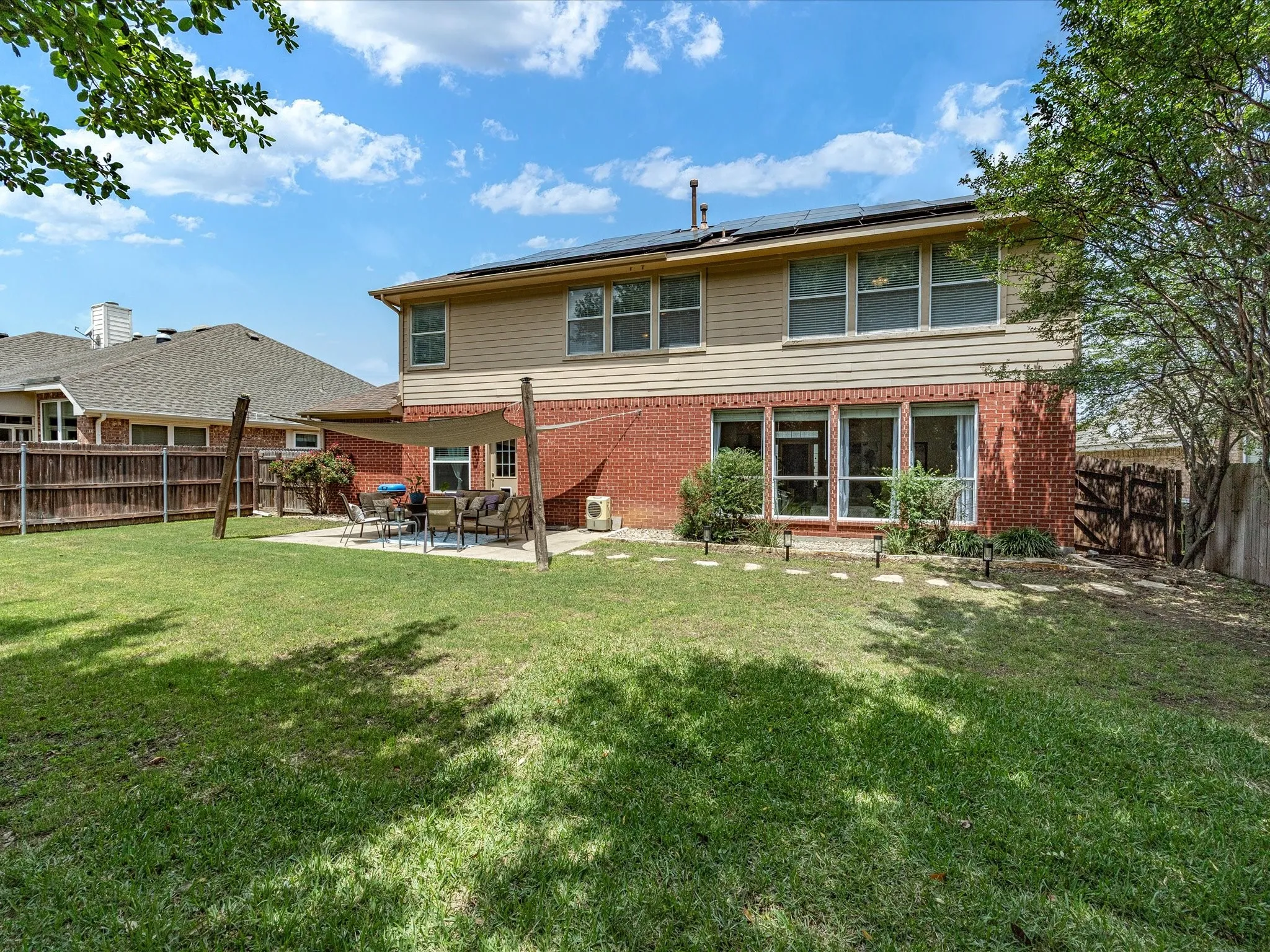 Expansive backyard featuring a large concrete patio, privacy fencing, solar panels on the roof, multiple rear windows, and exterior brick and siding finishes