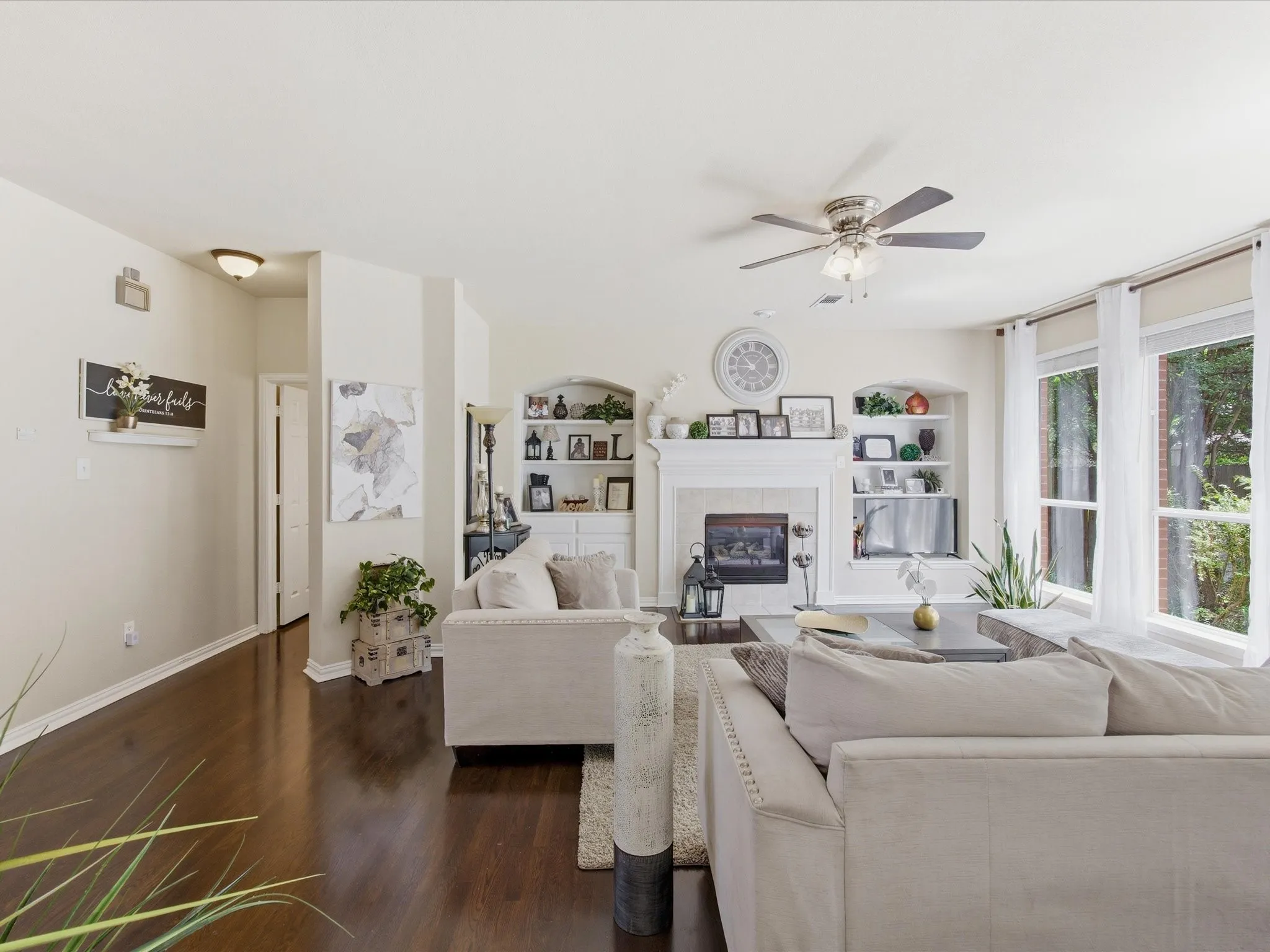 Living area featuring dark wood-finish flooring, a white mantel fireplace with tile surround, built-in shelving, a ceiling fan, and large windows with white trim