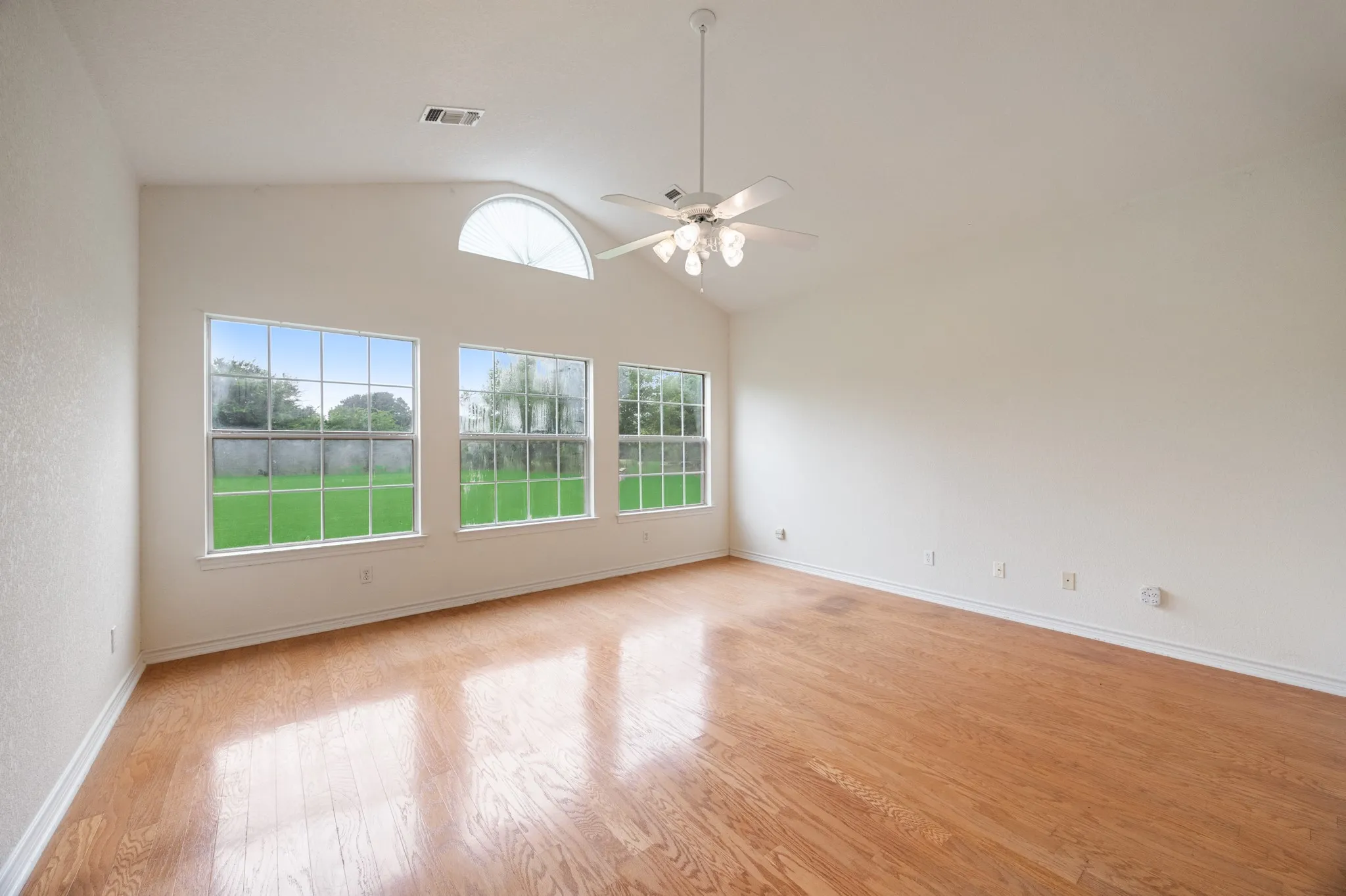 Spacious room featuring vaulted ceilings, wood-finish flooring, a semi-circular transom window, and three large windows overlooking a green lawn