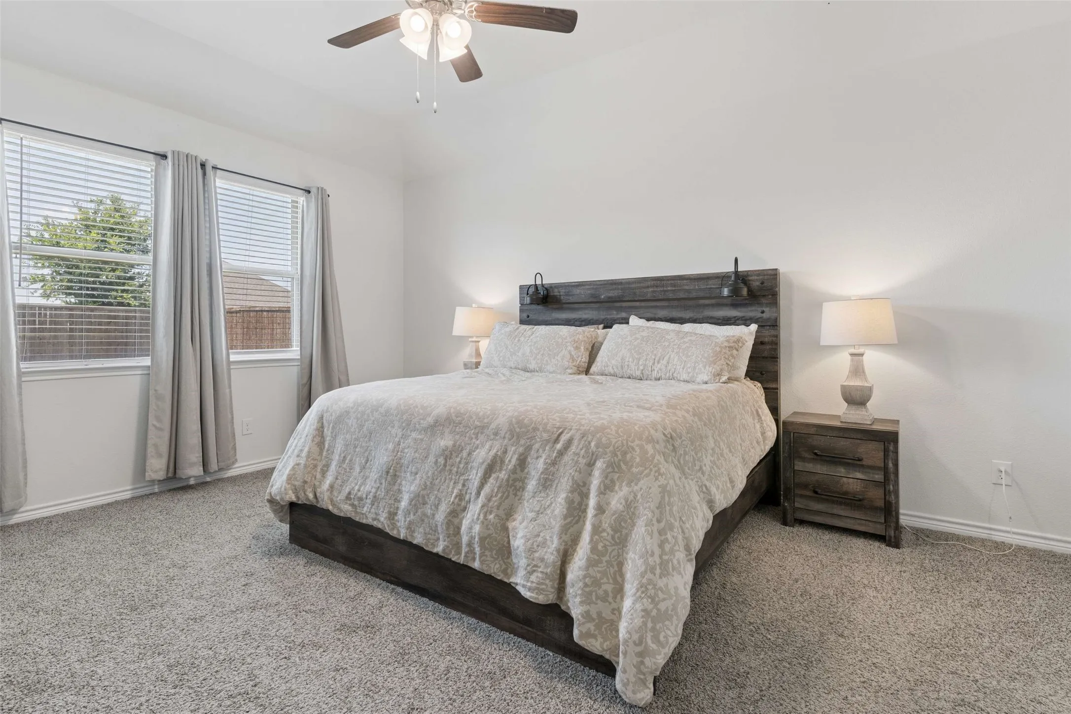 Bedroom featuring light gray carpeting, two large windows with blinds and drapes, and a ceiling fan with integrated lighting