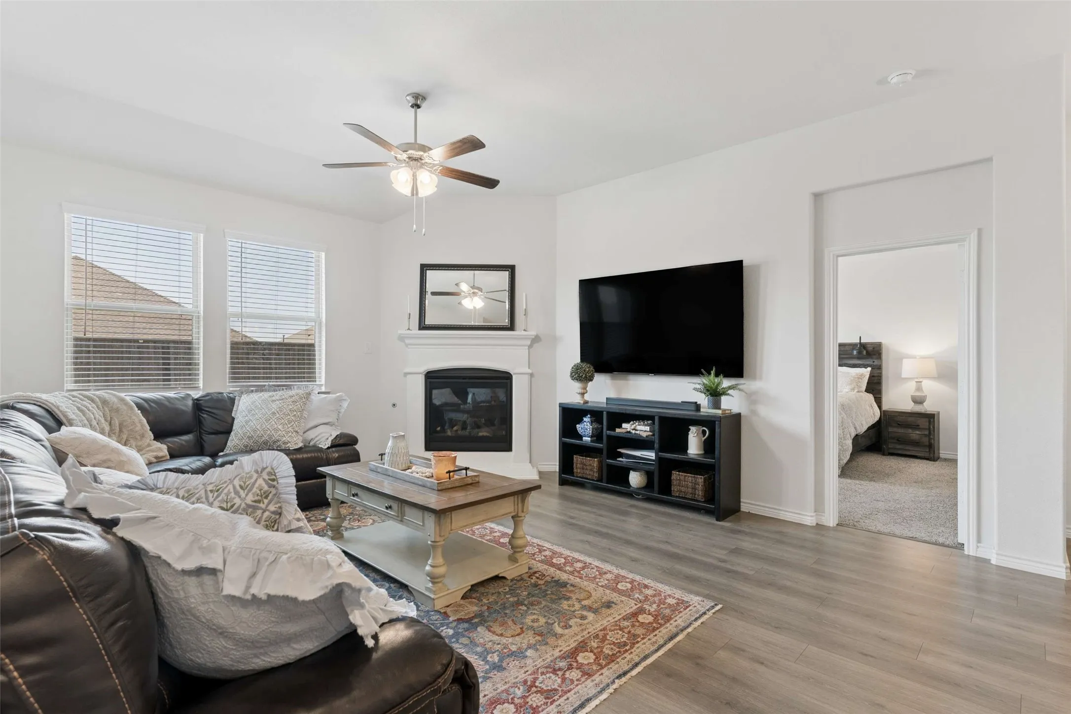Living area featuring wood-finish flooring, a white fireplace with mantel, ceiling fan with integrated lighting, and multiple windows