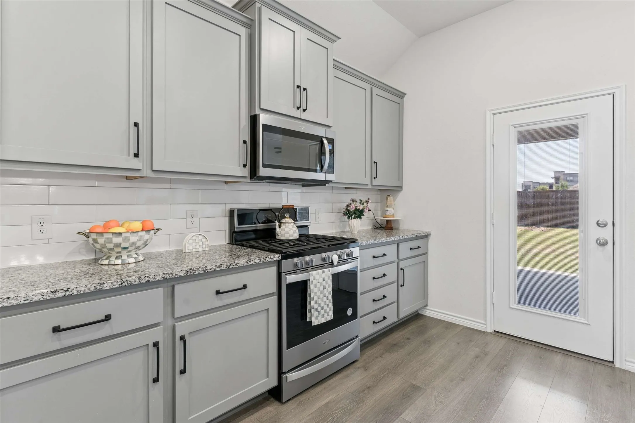Kitchen featuring grey cabinetry with matte black hardware, stainless steel appliances, speckled countertops, white subway tile backsplash, and wood-finish flooring