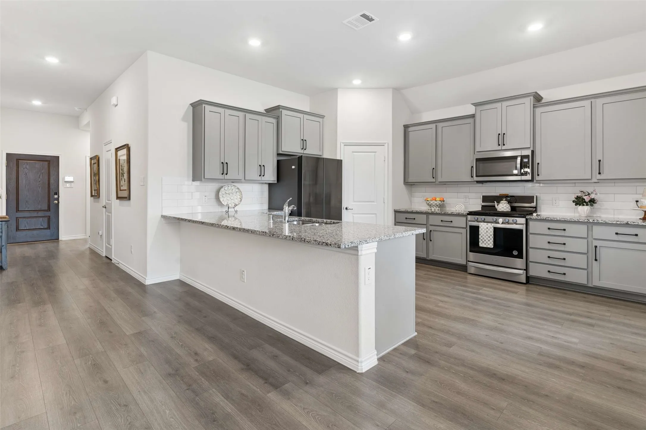 Open-concept kitchen featuring wood-finish flooring, light gray cabinetry, stainless steel appliances, speckled countertops, and a white subway tile backsplash