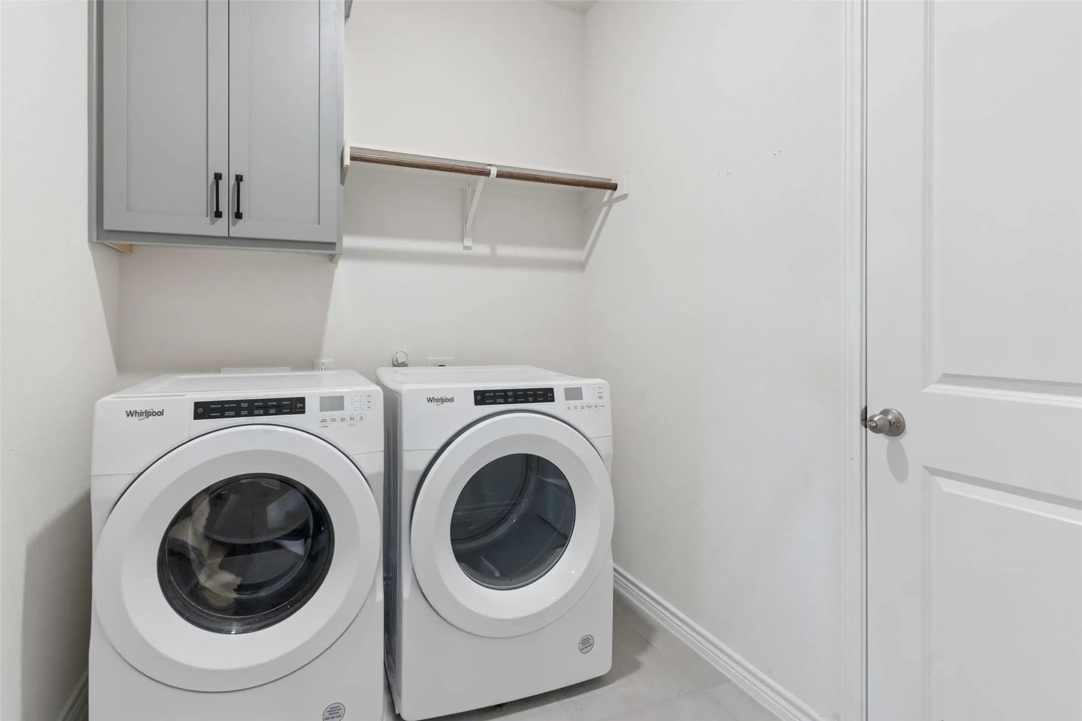 Dedicated laundry area featuring a light gray upper cabinet with matte black hardware, a wall-mounted shelving unit with a wood-finish rod, and neutral wall tones