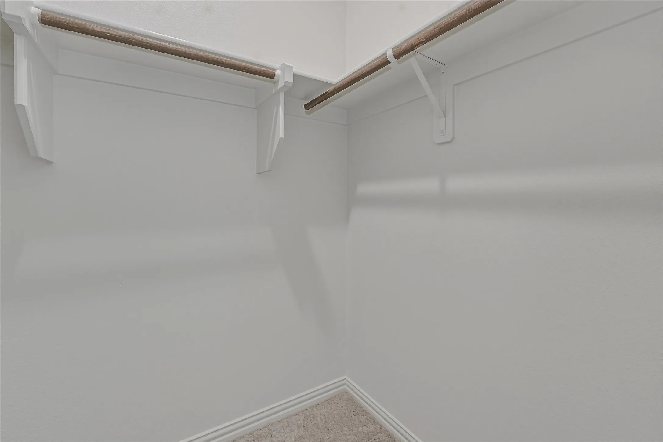 Corner closet with light-colored carpeting, white baseboards, and two wood-finish hanging rods supported by white shelving brackets