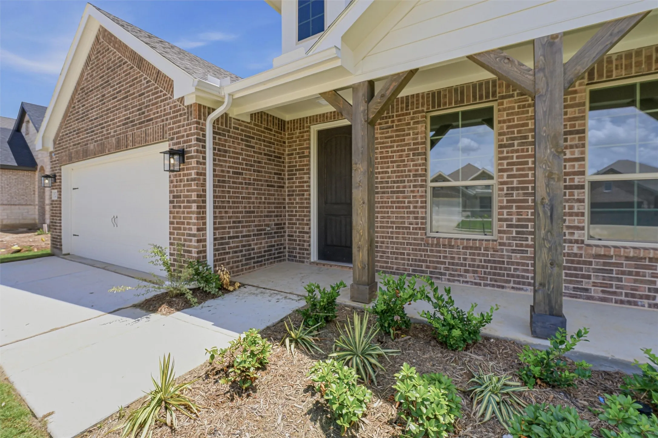 Brick facade with white trim, featuring a covered porch with natural wood columns, a white garage door, and landscaping elements