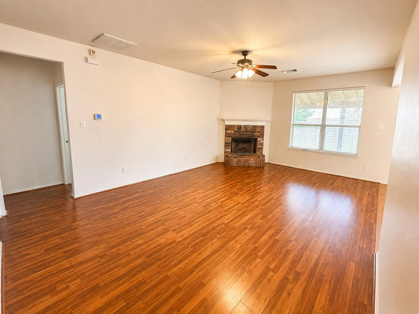 Wood-finish flooring throughout, brick fireplace with hearth, ceiling fan, and large window with blinds