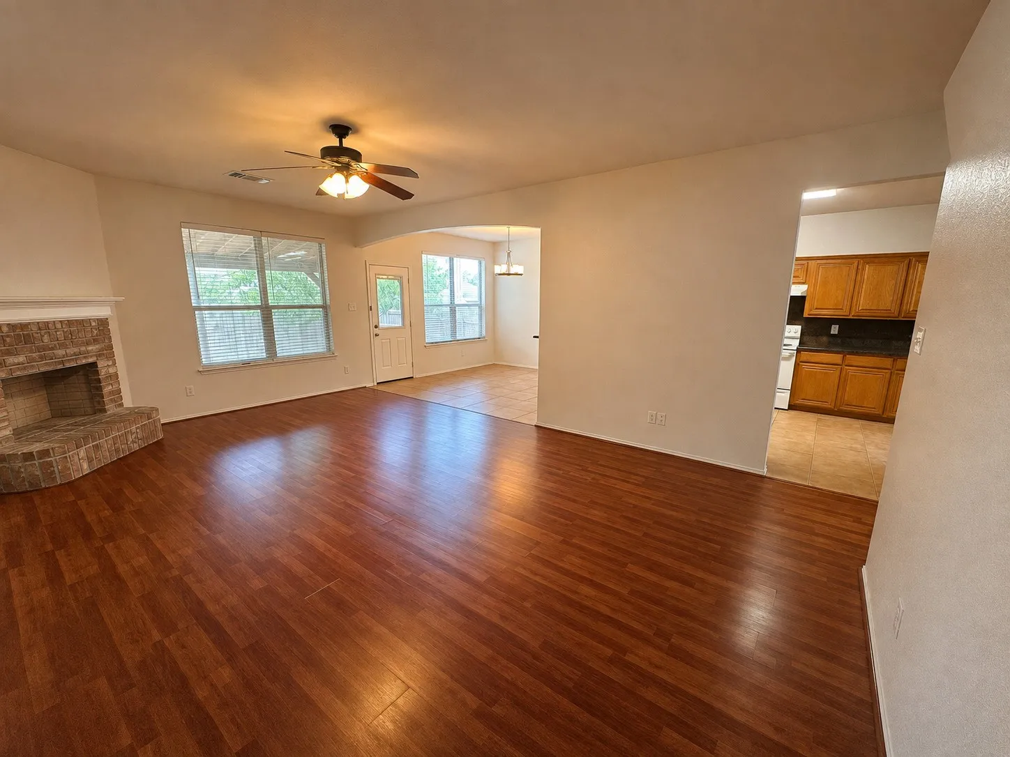 Wood-finish flooring throughout, brick fireplace with mantel, ceiling fan with integrated lighting, multiple windows, and an arched entryway