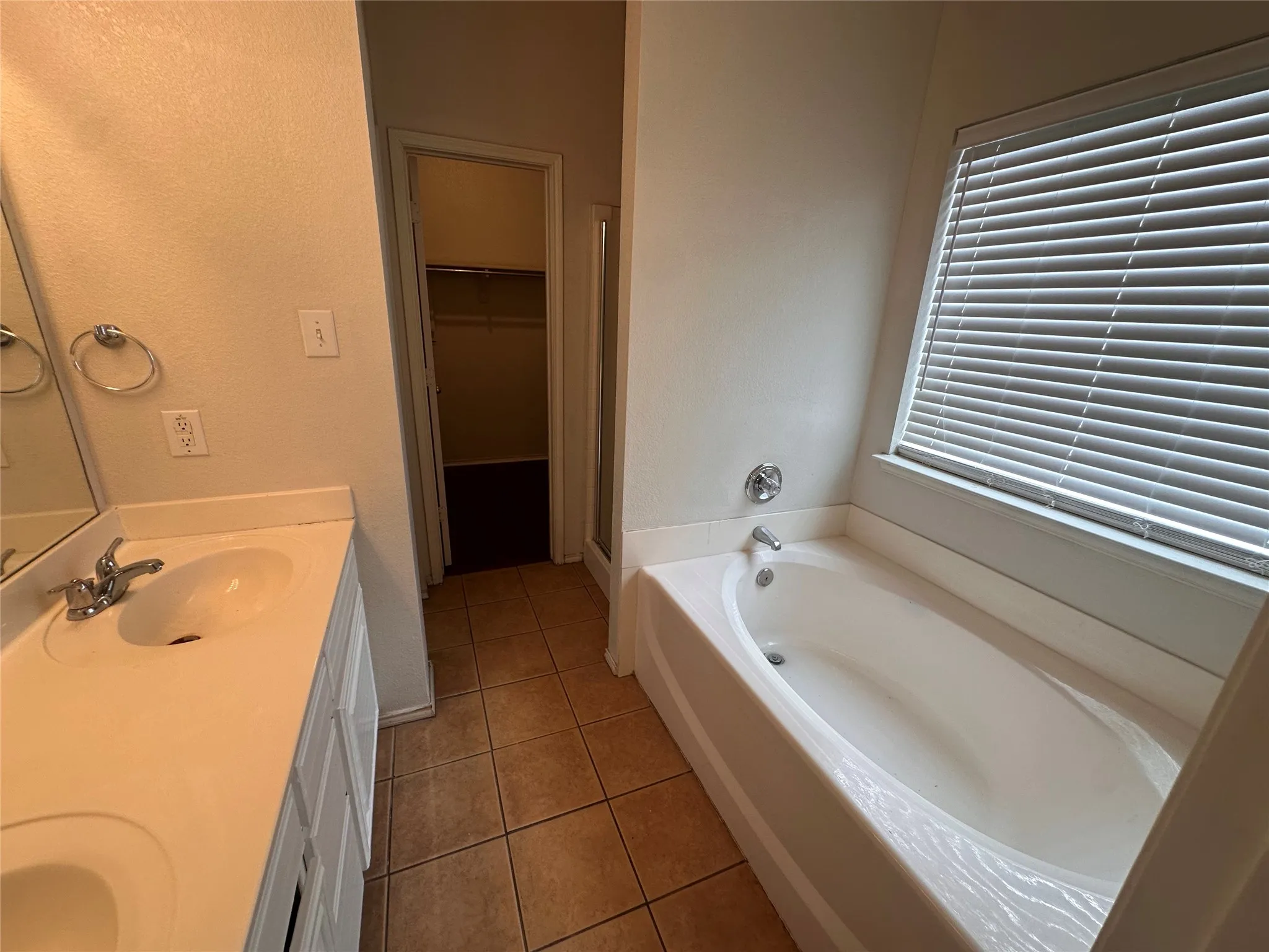 Spacious primary bathroom featuring dual vanities with ceramic sinks, tile flooring, an alcove soaking tub with jets, and horizontal blinds at the window for privacy and light control