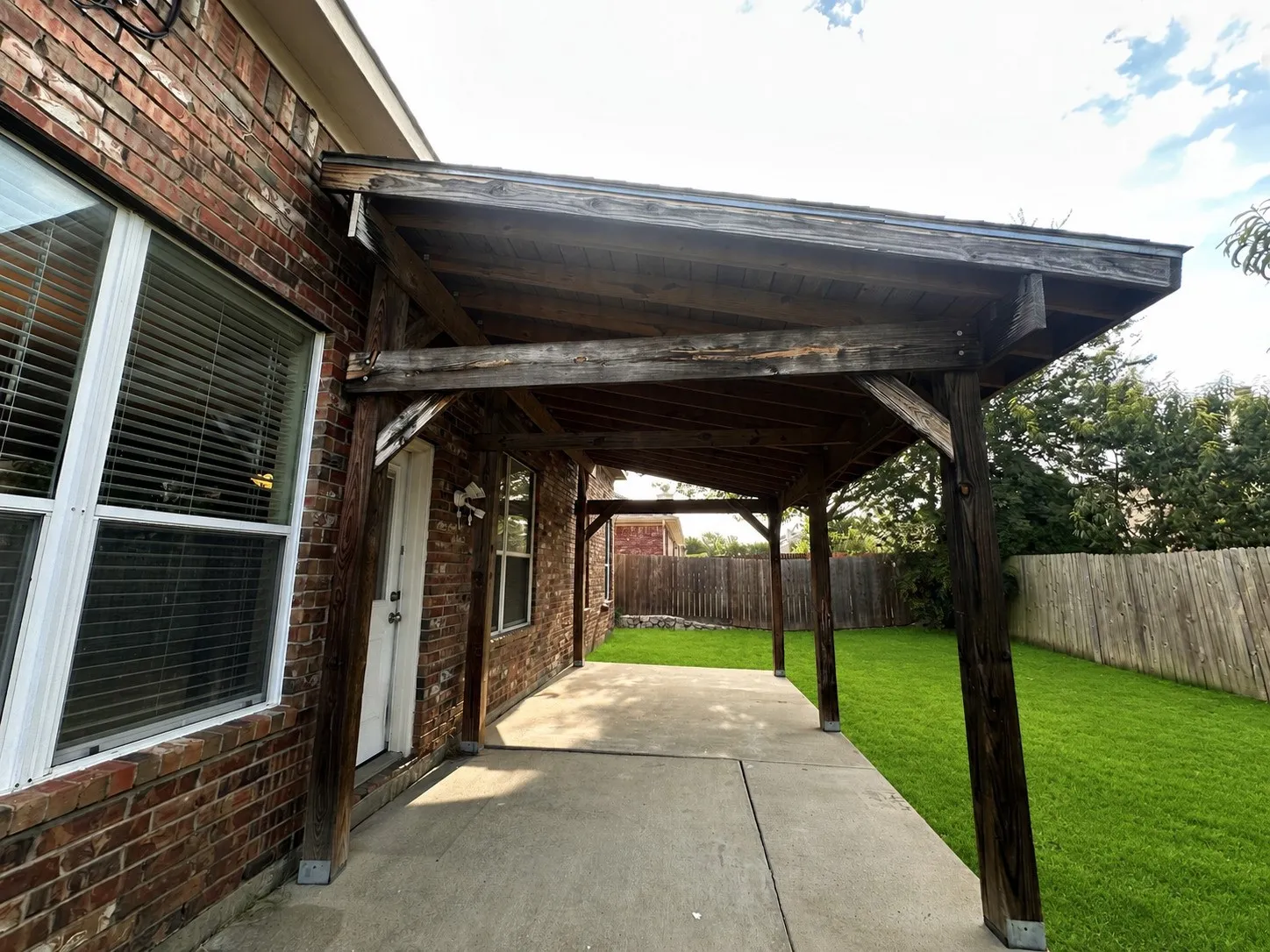 Expansive concrete patio with a wood-frame covered area, brick exterior, white-frame windows, and a manicured lawn