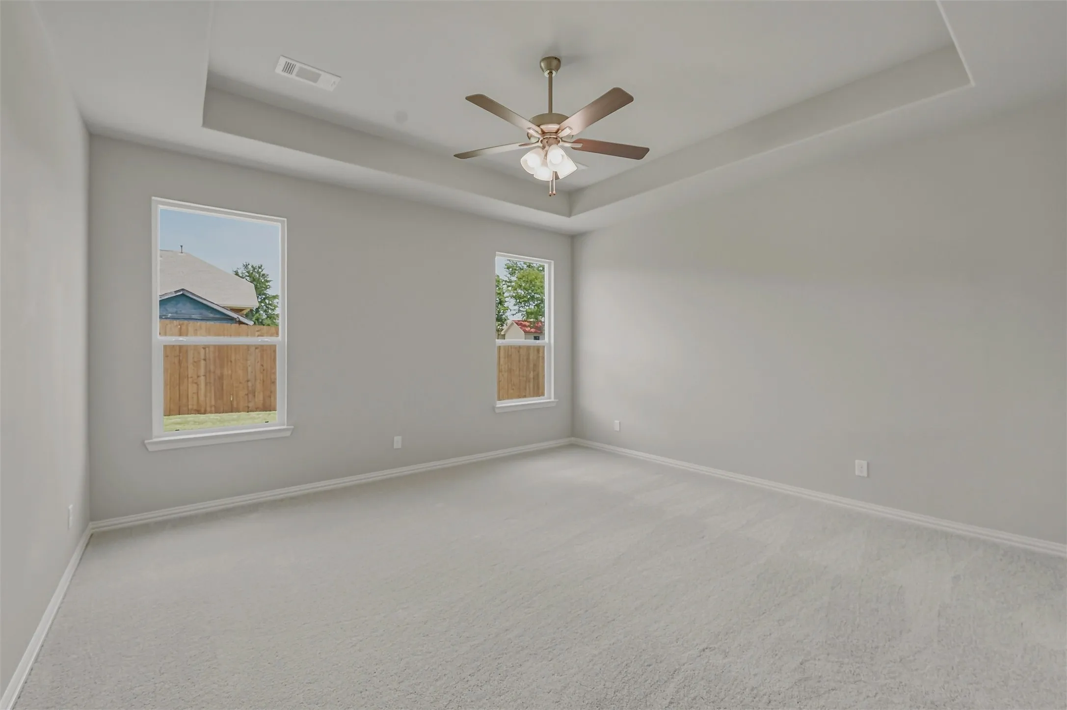 Spacious room featuring light gray walls, plush gray carpeting, and a tray ceiling with a ceiling fan light fixture