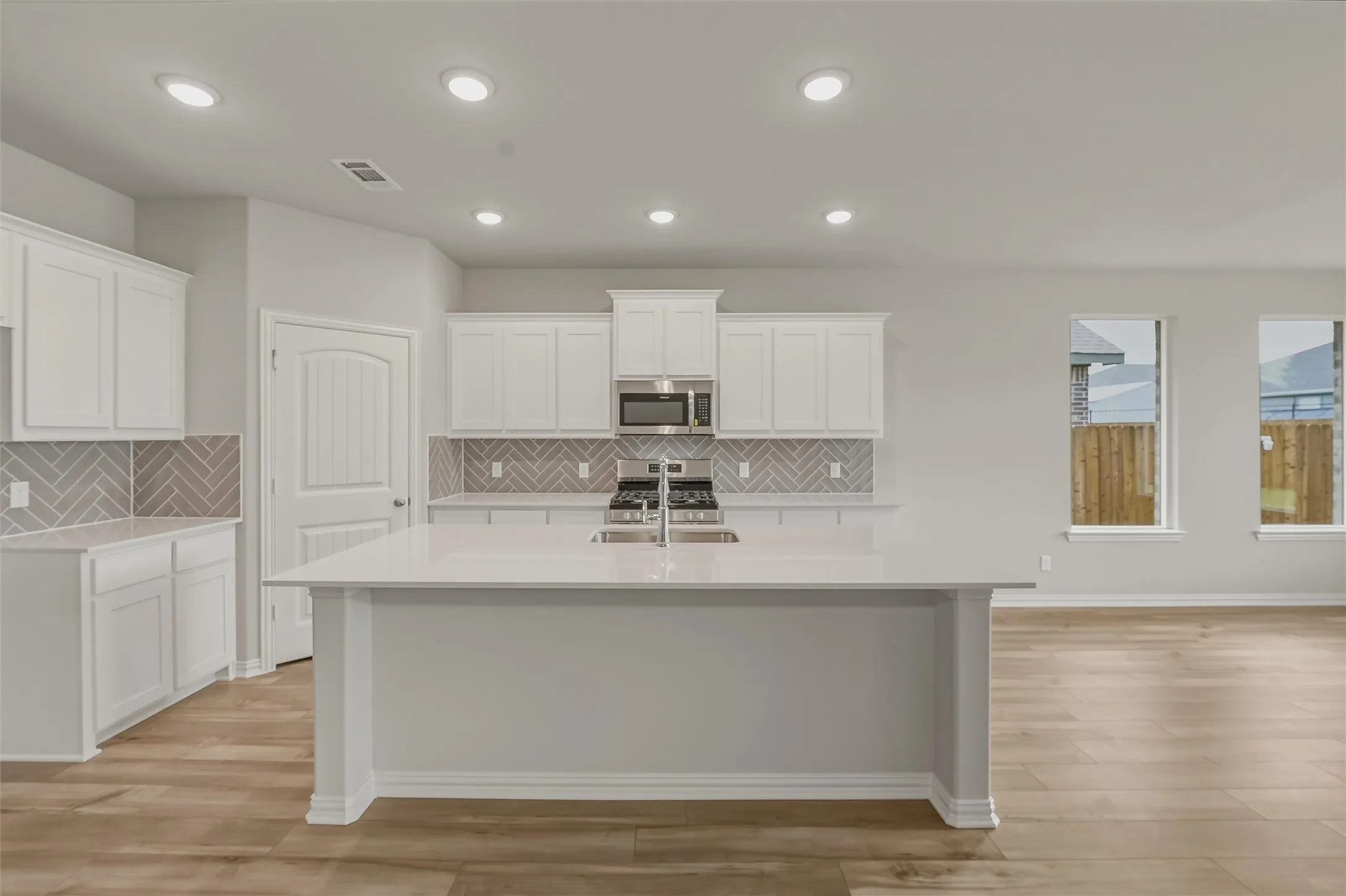 Modern kitchen featuring white shaker cabinetry, light-toned countertops, a herringbone tile backsplash, an island with an integrated sink, and wood-finish flooring