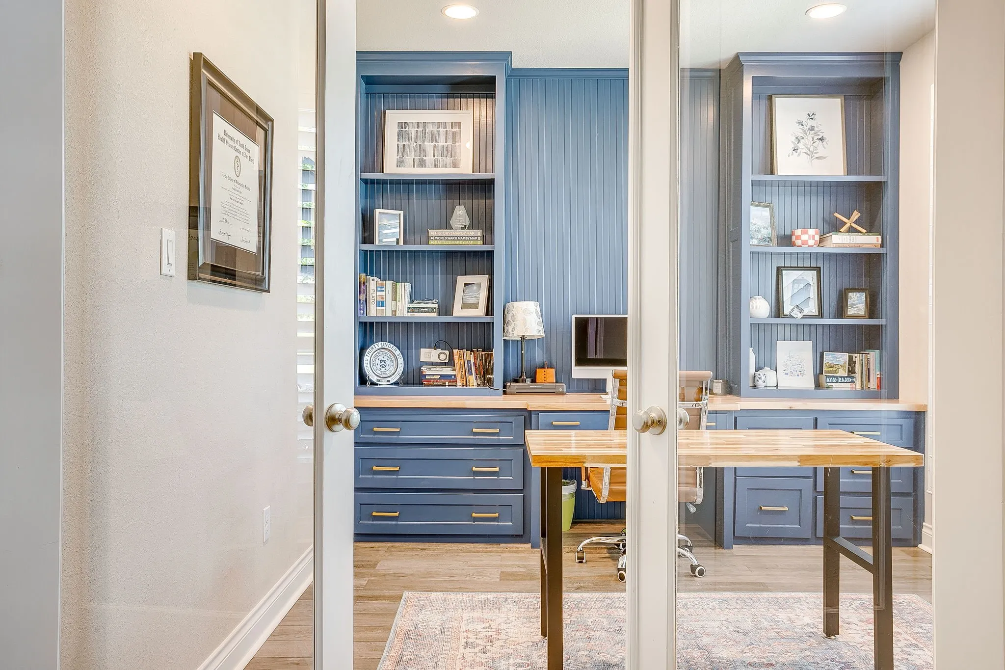 Located at the front of the home, double glass doors open to a stunning office space featuring built-in blue cabinetry, integrated shelving, beadboard accents, and light wood-finish countertops.
