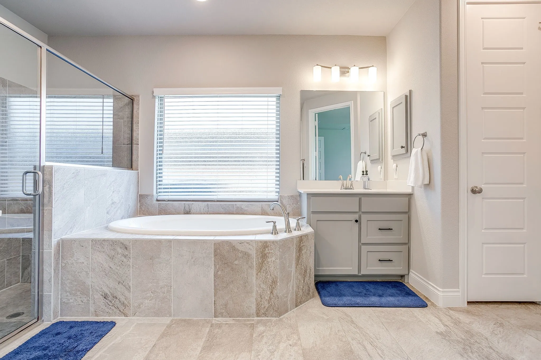 Relax in this spacious primary bathroom featuring a soaking tub with surrounding stone-look tile, an adjacent glass-enclosed shower, double vanities, and separate his-and-hers walk-in closets.
