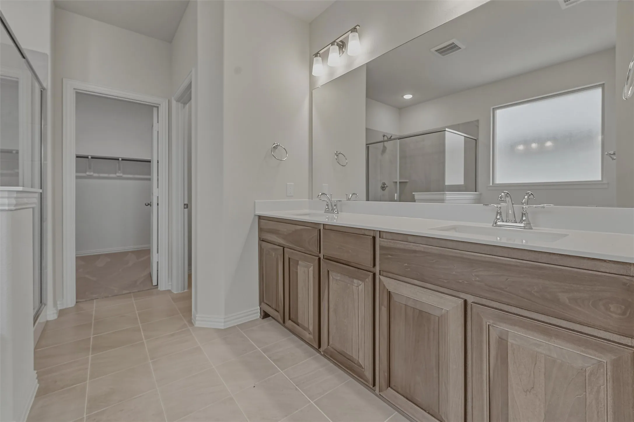 Spacious bathroom featuring a dual vanity with light-colored wood-finish cabinetry, a large frameless mirror, and a frosted privacy window