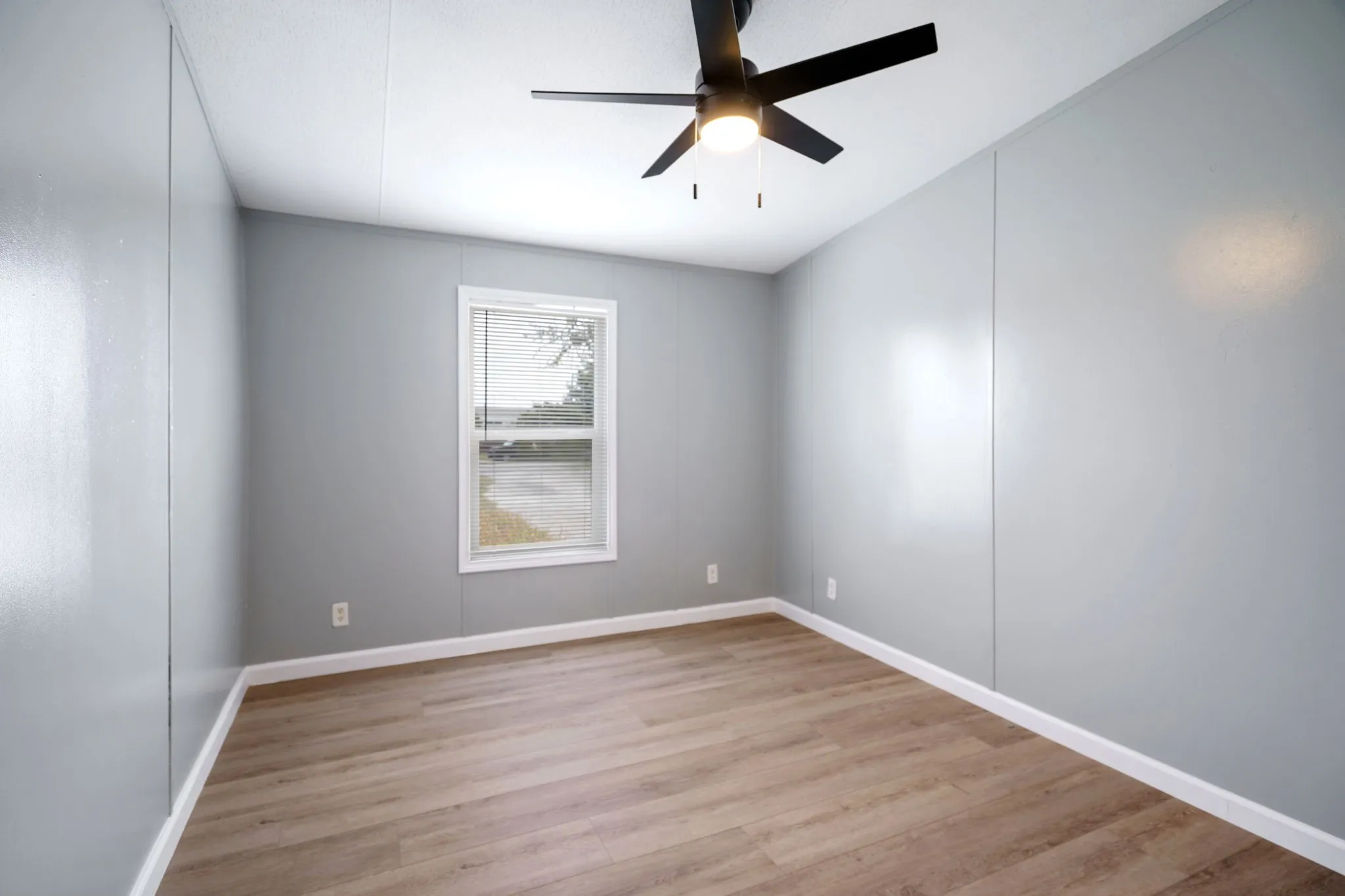 Interior space featuring wood-finish flooring, light gray walls, a white-framed window with blinds, a white ceiling, and a black ceiling fan with an integrated light