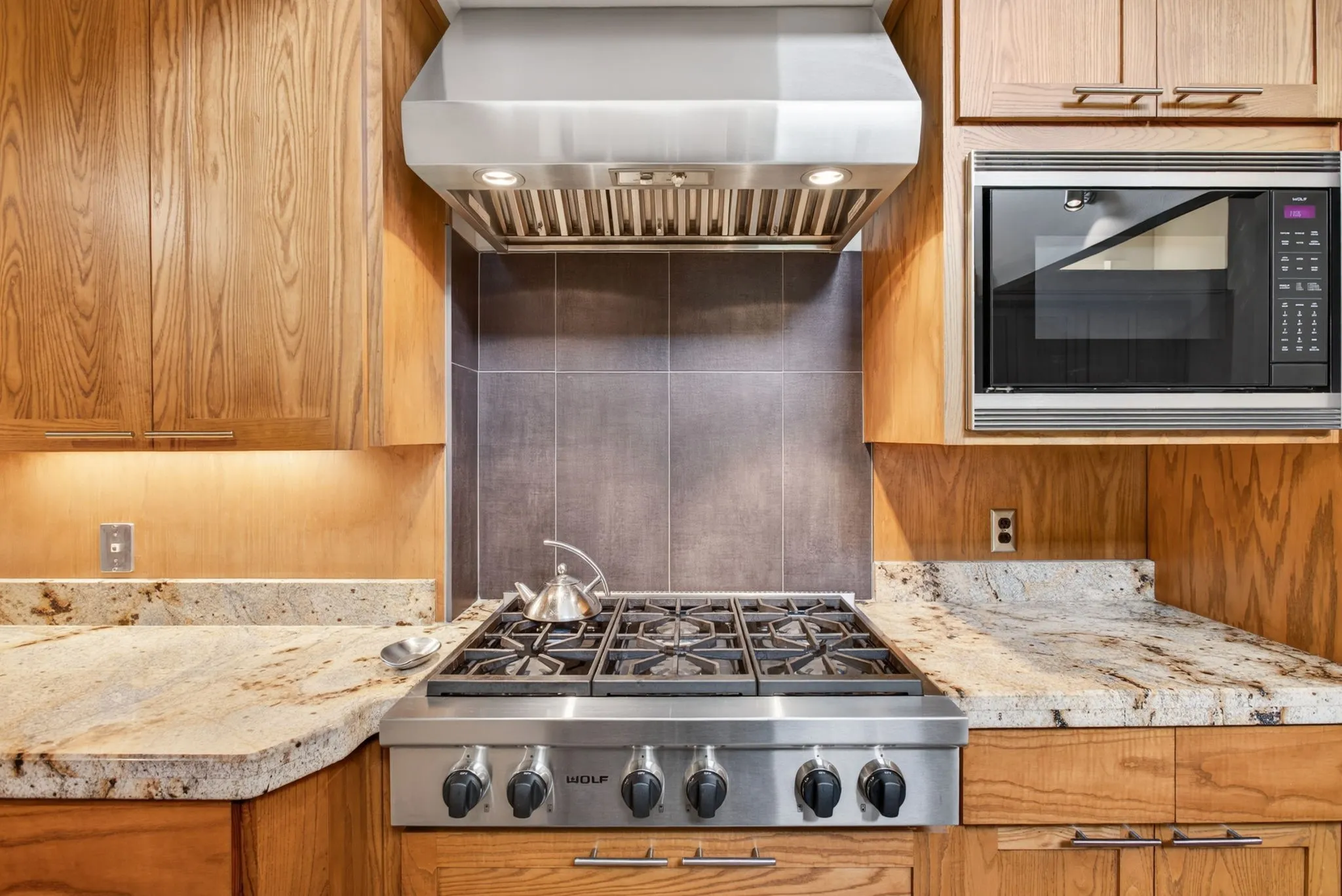 Stainless steel range with a professional-grade hood, natural wood cabinetry, stone countertops, integrated microwave, and a tiled backsplash