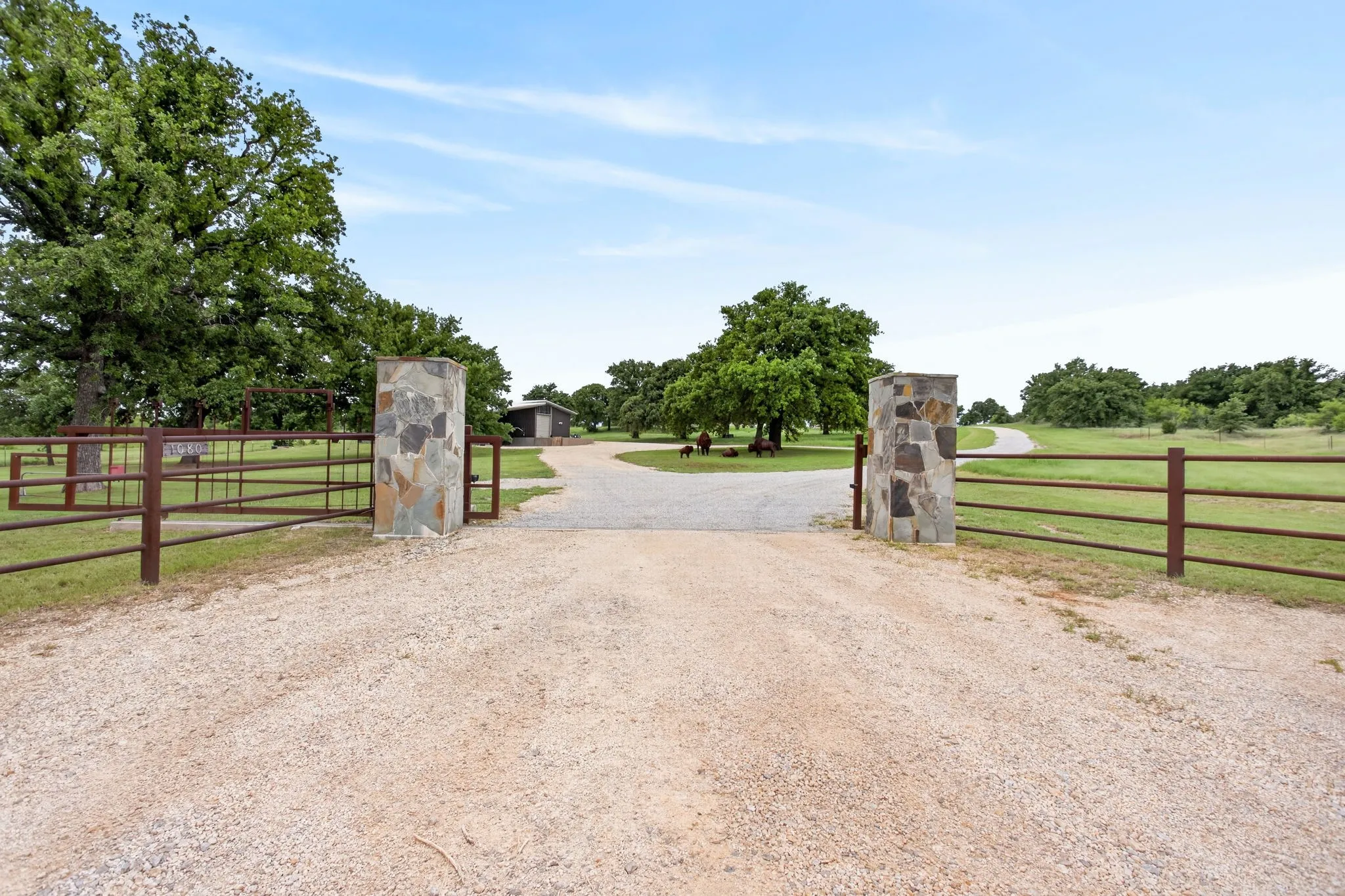 Gravel driveway entry framed by stone pillars and a metal gate, opening to a sprawling property with mature trees and a distant outbuilding