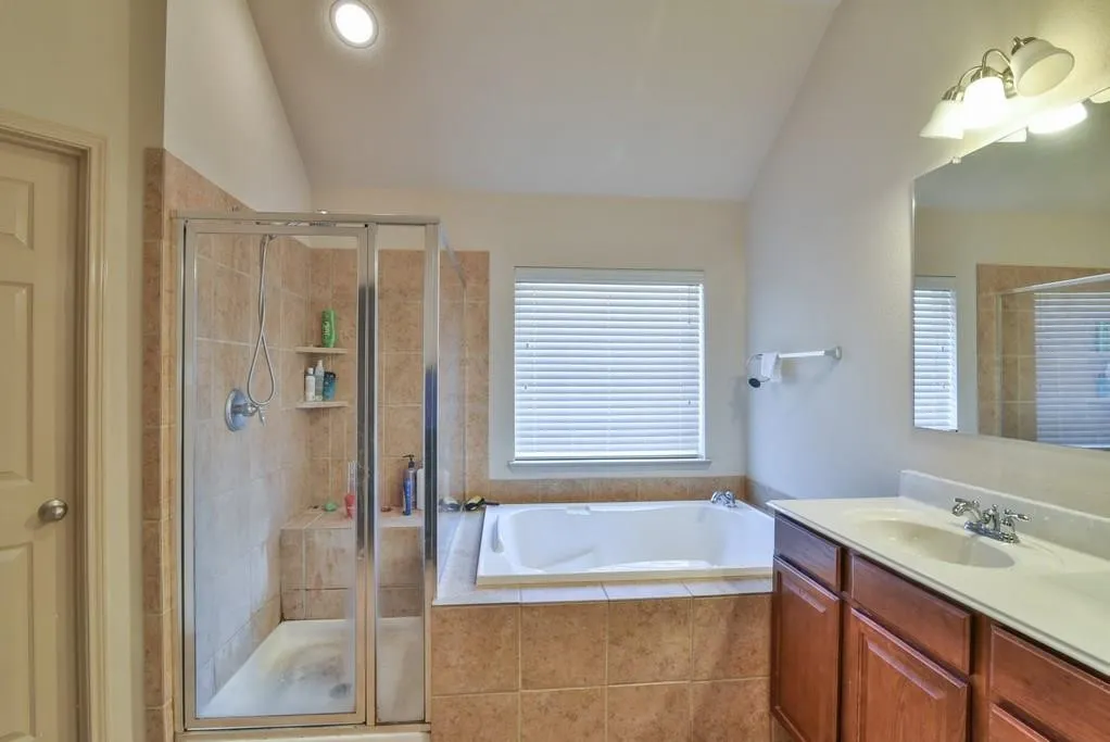 Bathroom featuring a built-in tub with tile surround, a separate glass-enclosed shower with tile walls and integrated shelving, a wood-finish vanity with a single sink and light-toned countertop, and recessed ceiling lighting