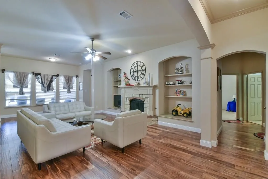 Spacious living area featuring wood-finish flooring, a stone-surround fireplace, built-in shelving, arched doorways, and a ceiling fan