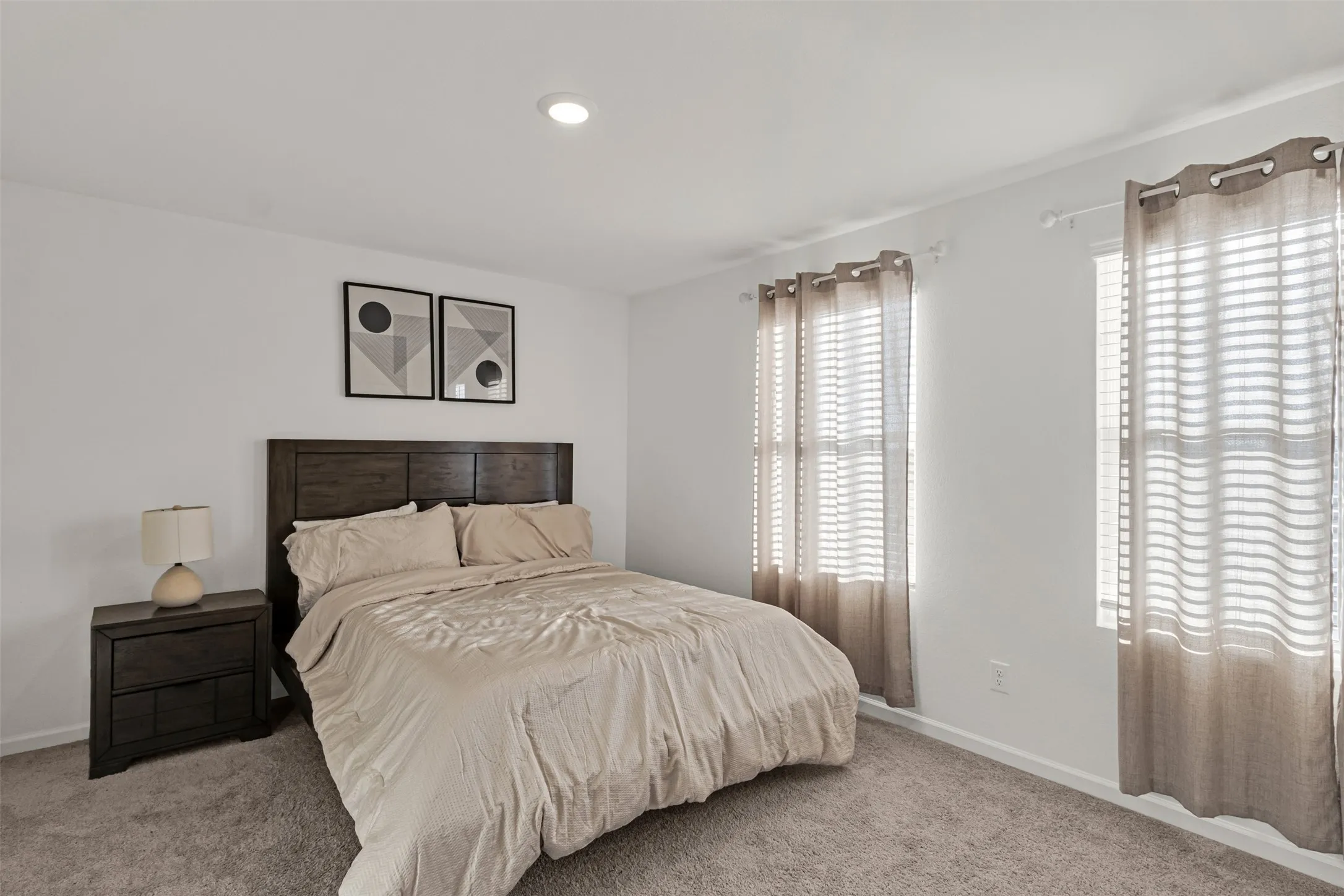 Carpeted room featuring a dark wood-finish headboard and nightstand, recessed ceiling lighting, and two windows with blinds and curtain rods