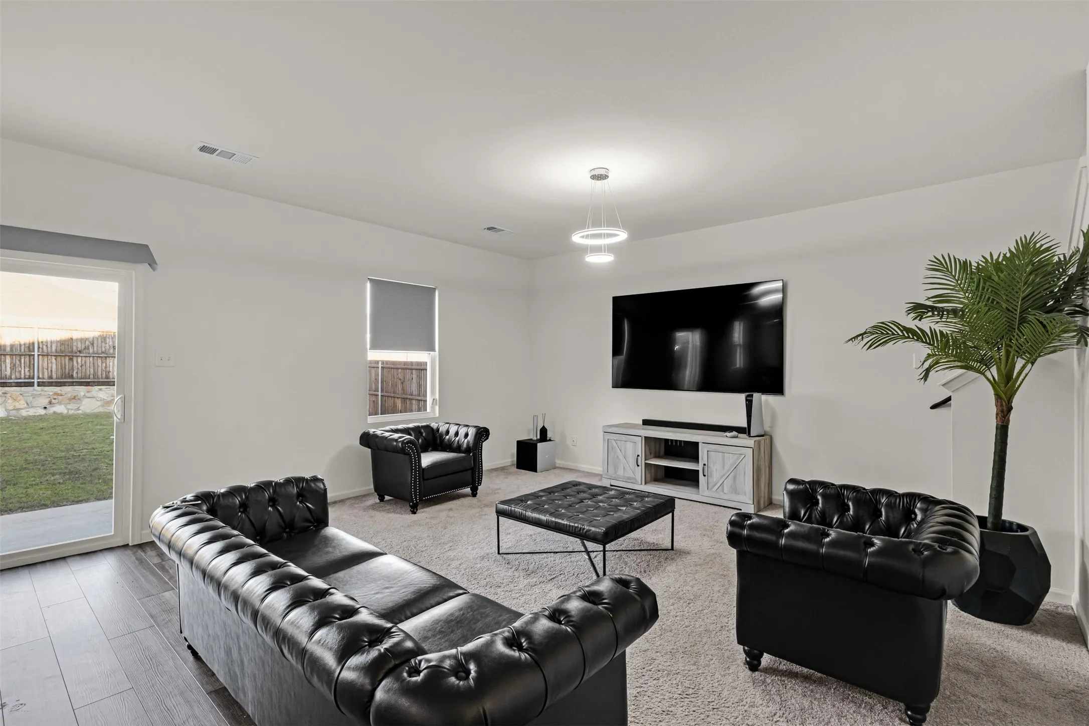 Spacious living area featuring light-colored carpeting, a sliding glass door to exterior, a single window with a roller shade, and modern ceiling lighting