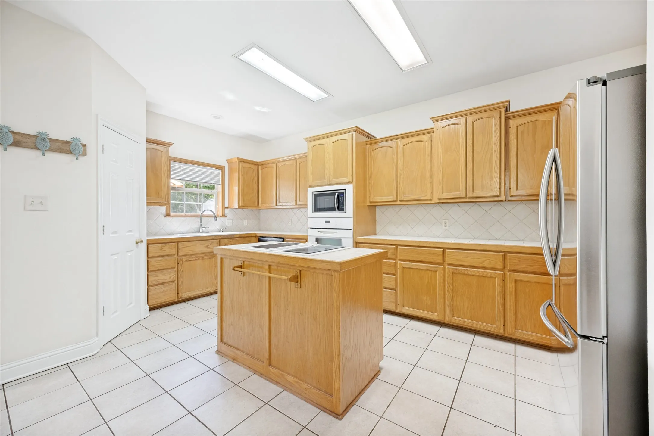 Kitchen featuring abundant wood cabinetry, an island with a built-in cooktop, white tile flooring, a built-in microwave, and a stainless steel refrigerator