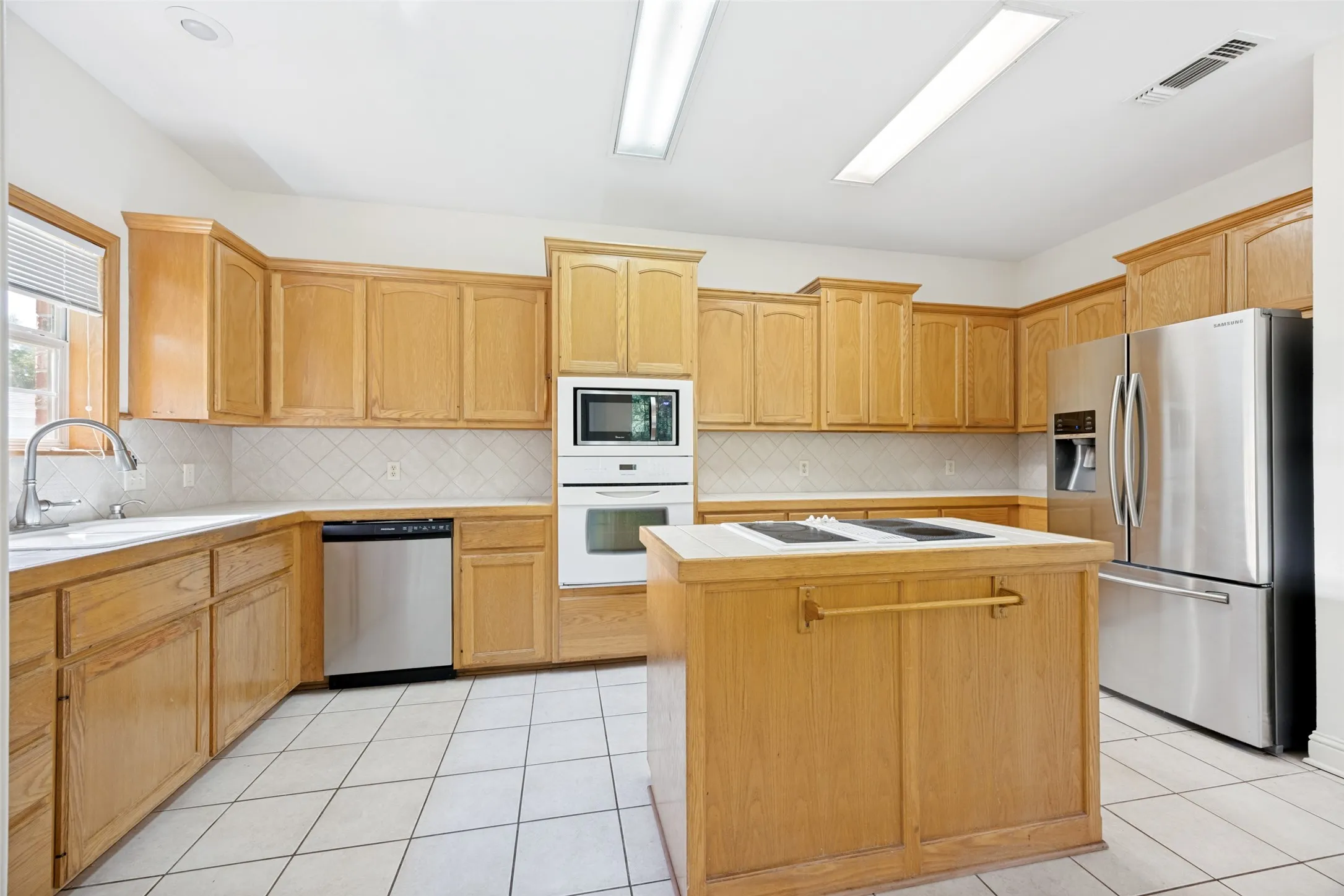 Bright kitchen featuring extensive light wood cabinetry, a central island with a cooktop, built-in stainless steel appliances, a tile backsplash, and ceramic tile flooring