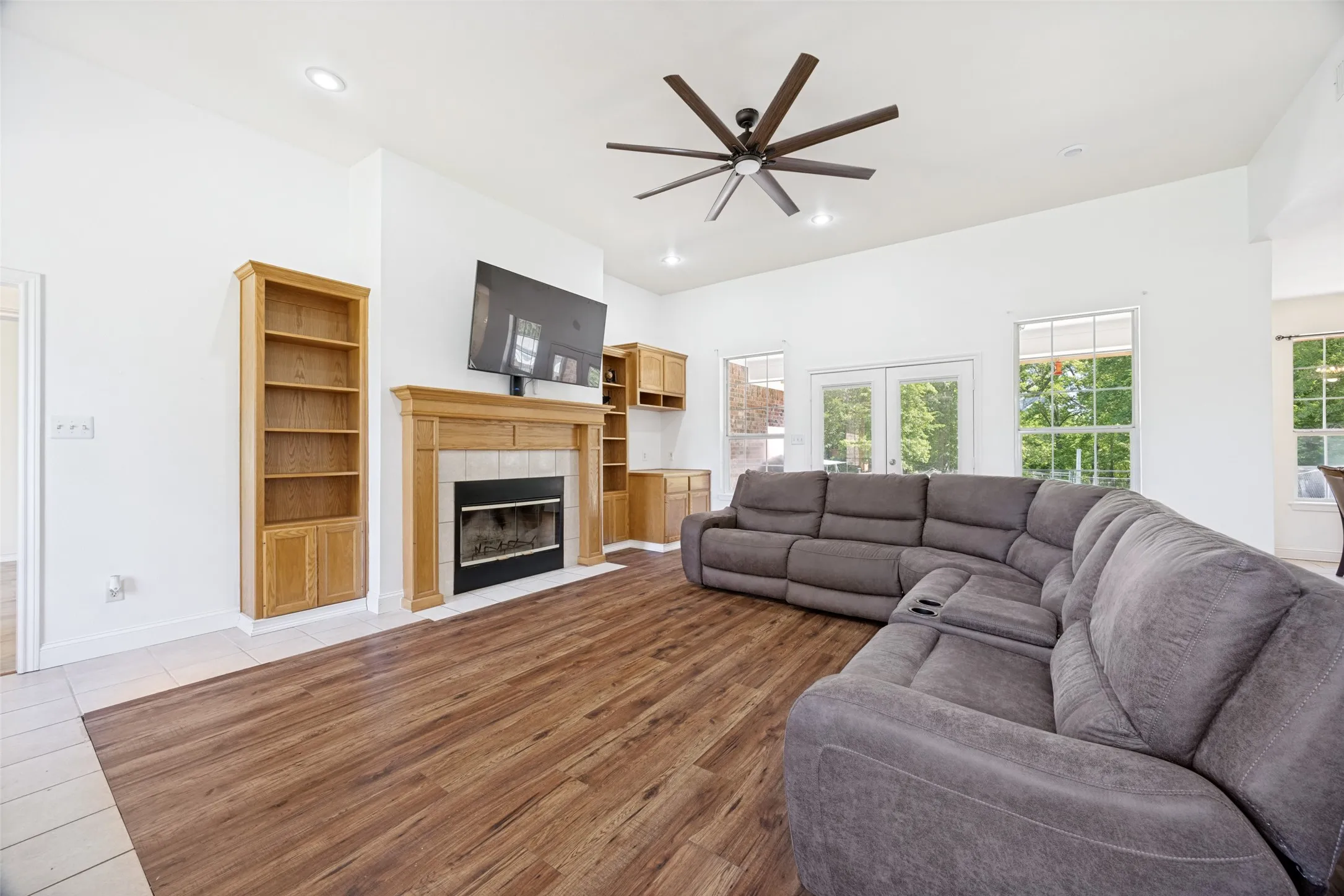 Bright living space featuring wood-finish flooring, a tile fireplace surround with a wood mantel, built-in wooden shelving, a multi-blade ceiling fan, and French doors leading outdoors
