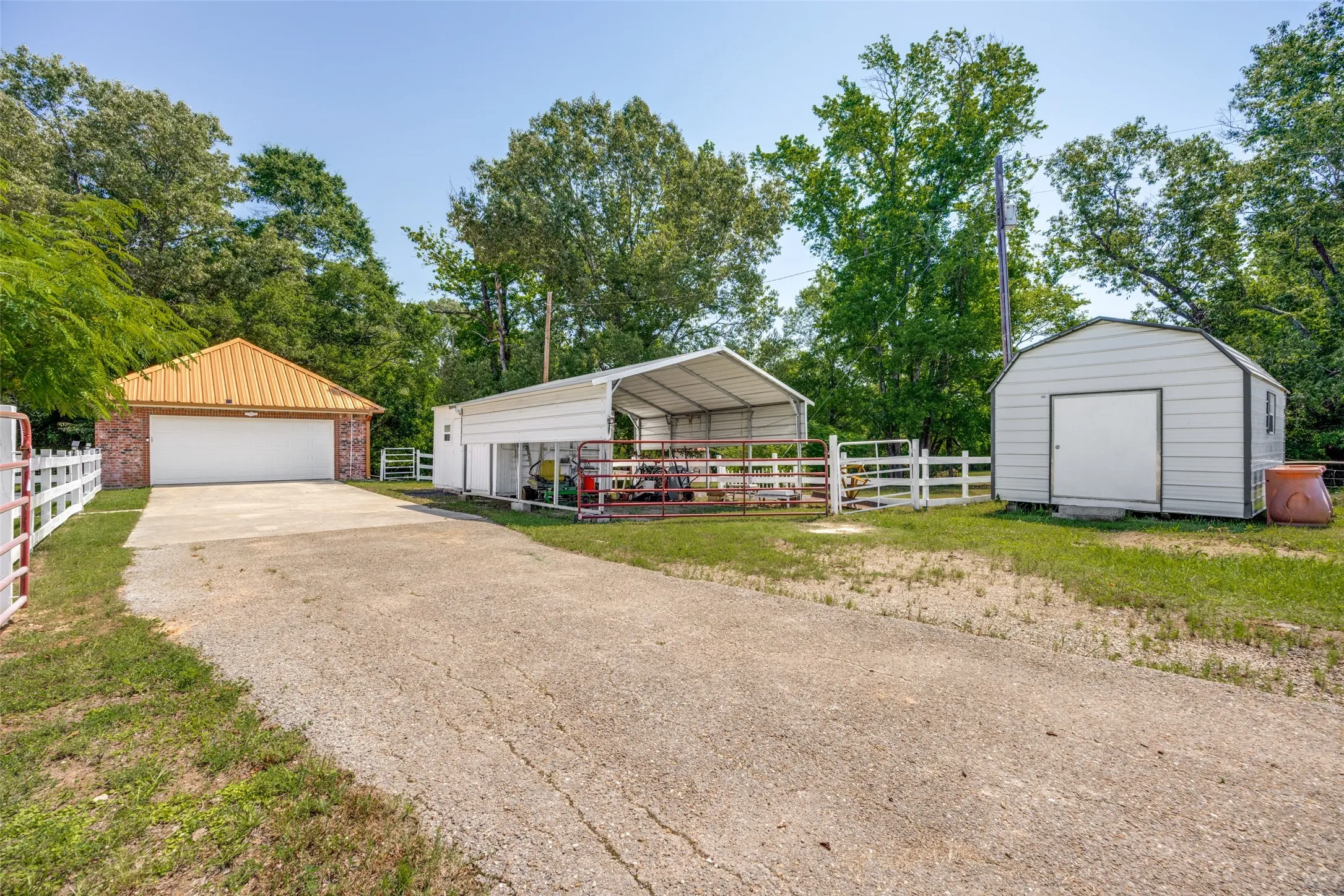 Concrete driveway leading to a detached brick garage with a metal roof, white garage door, white fencing, a metal carport, and a separate storage shed