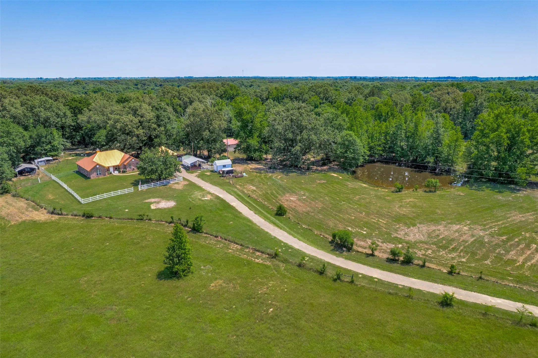 Expansive property featuring a brick residence with a light-colored roof, a lengthy driveway, and a pond