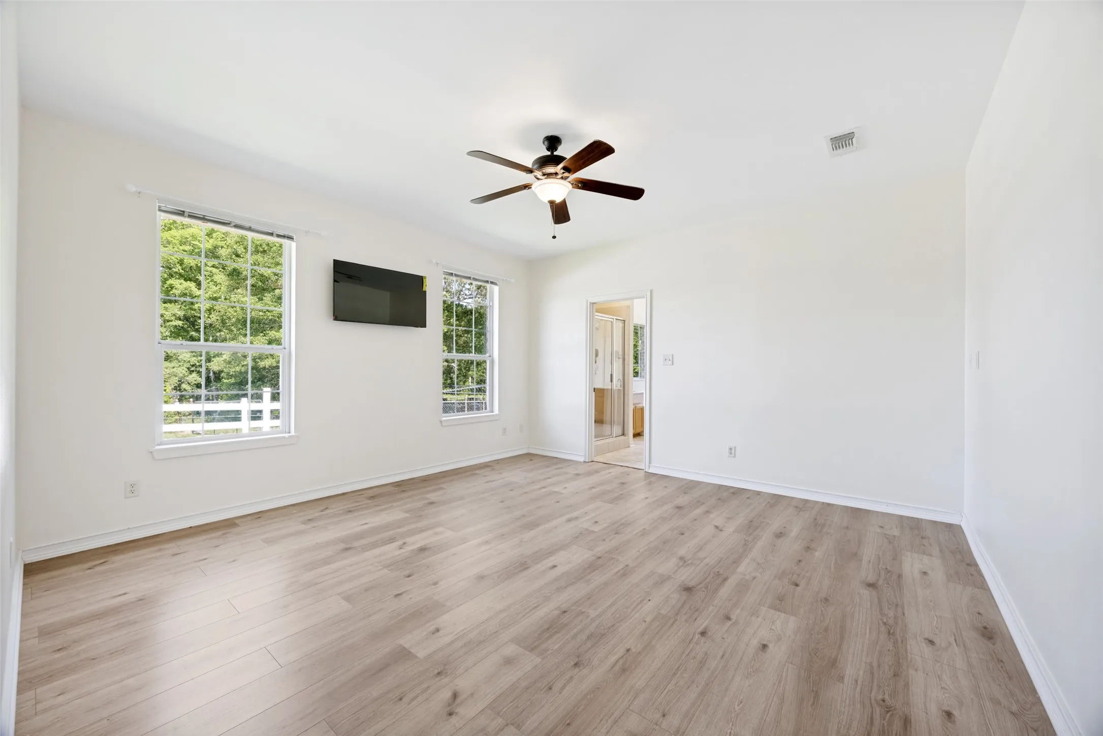 Spacious room with light wood-finish flooring, crisp white walls, two large windows, and a ceiling fan with integrated lighting