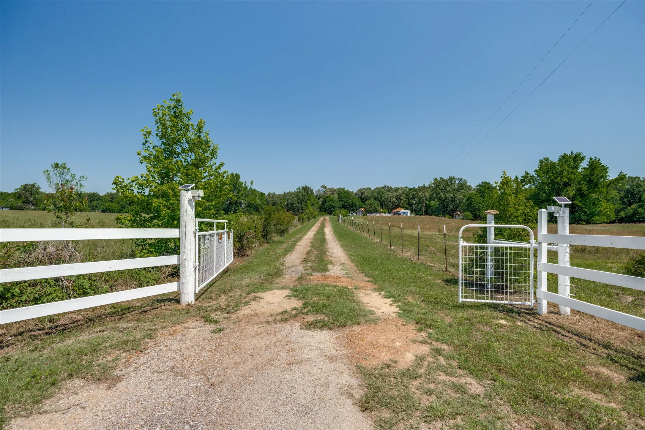 Gated entry featuring a white board fence, solar-powered gate openers, and a gravel driveway extending into the property