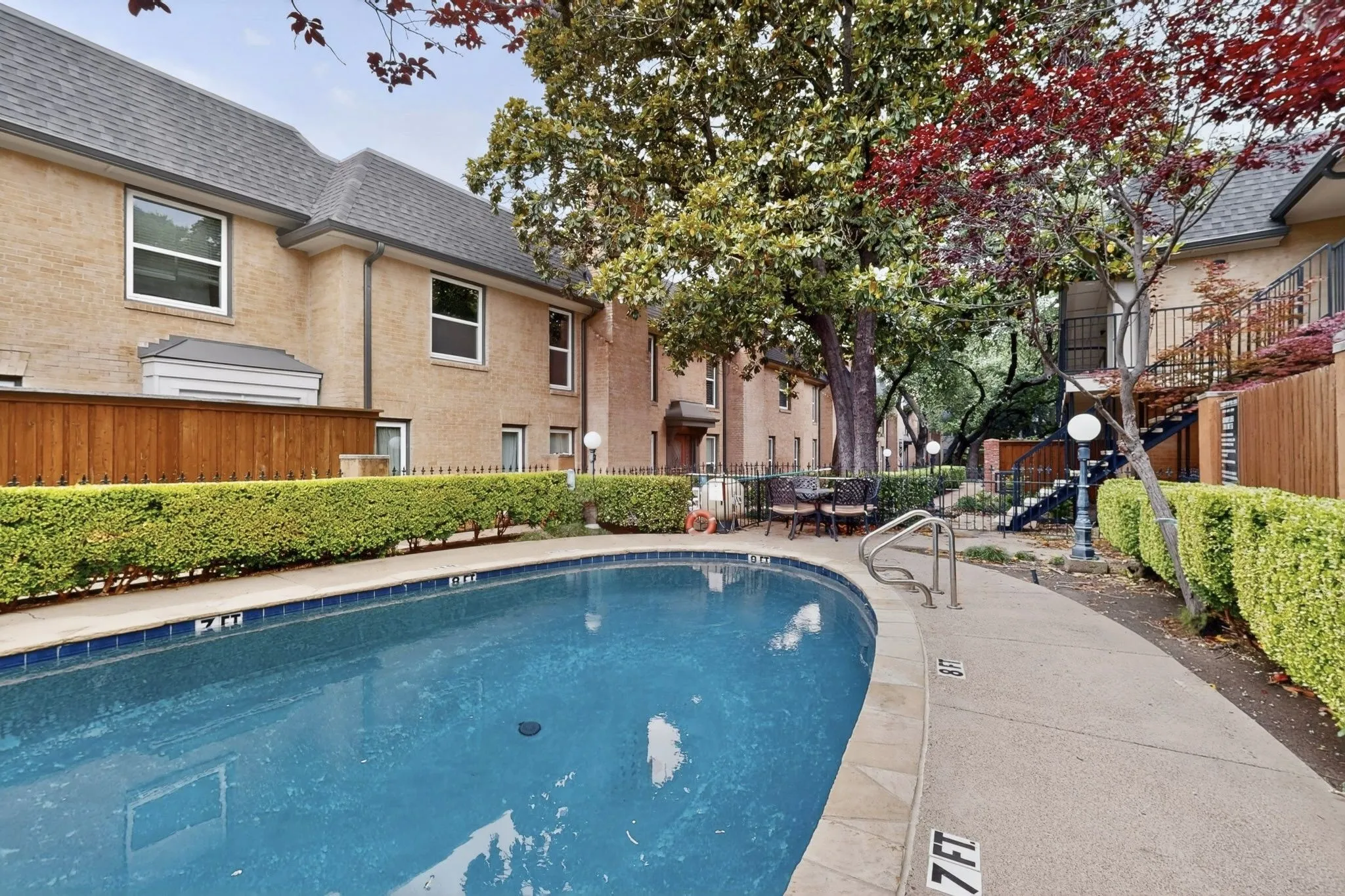 Curved swimming pool with concrete decking, surrounded by manicured hedges and a privacy fence