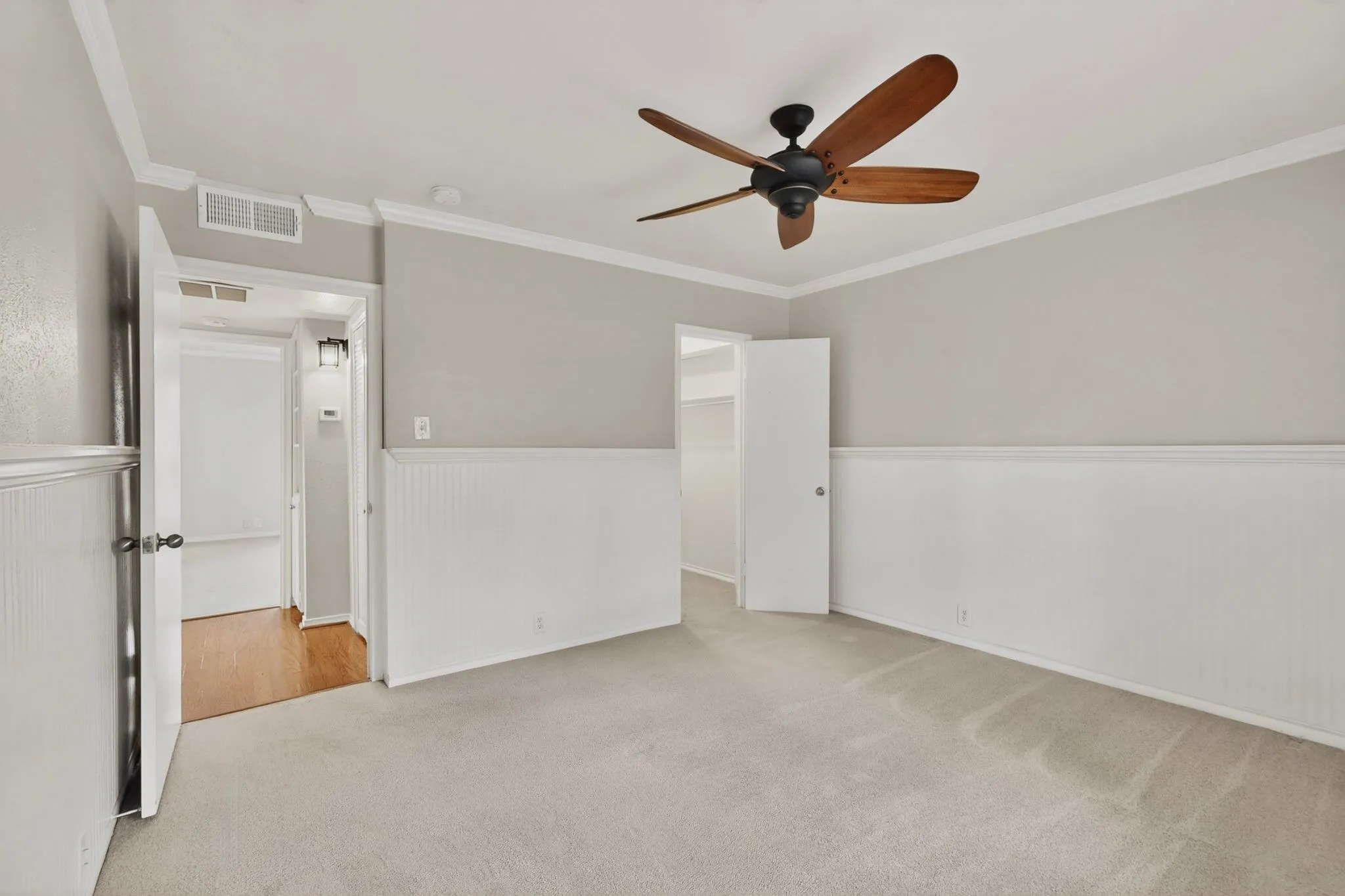 Spacious room featuring neutral carpeting, light gray painted walls, white wainscoting, and a ceiling fan with wood-finish blades
