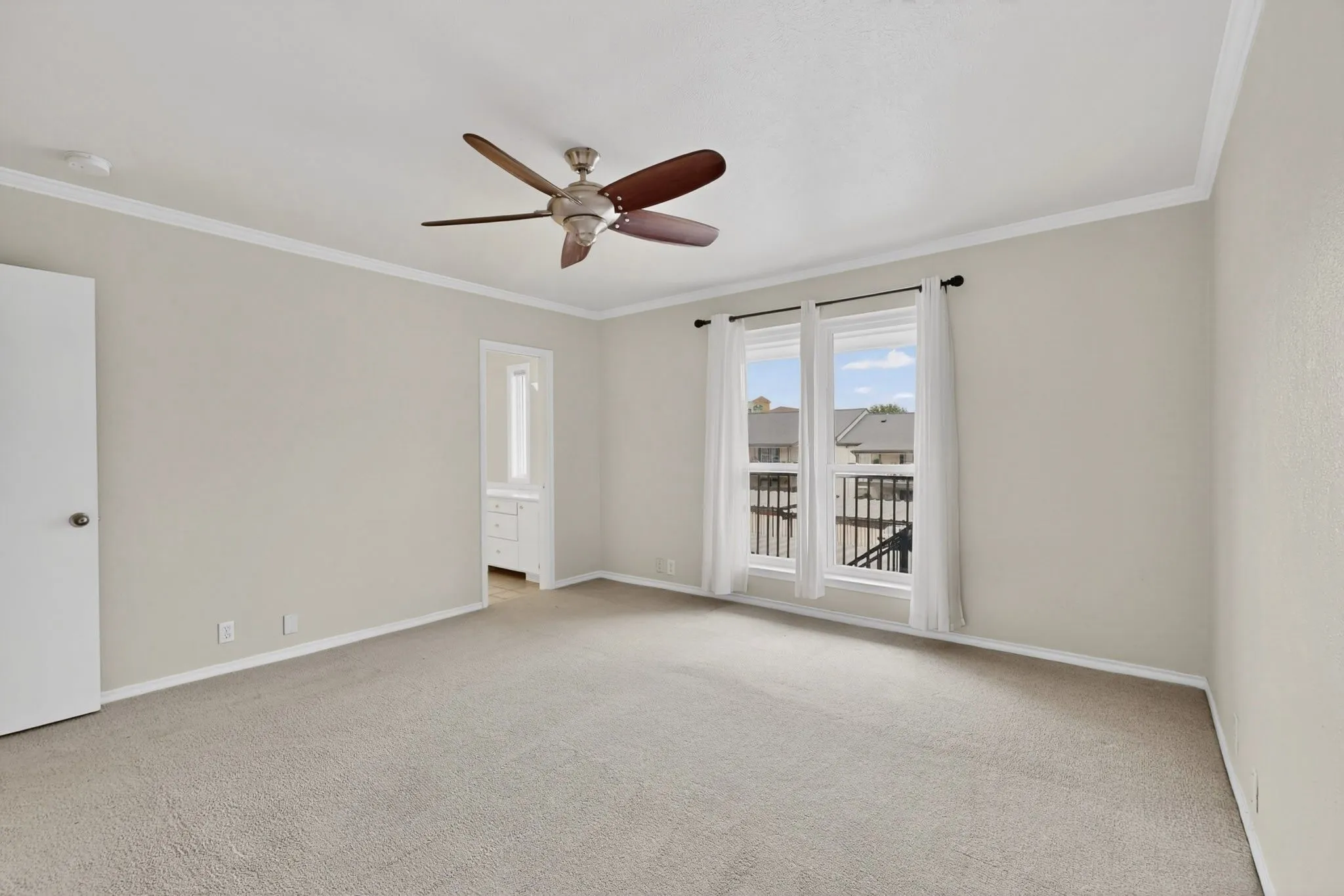 Spacious room with light-colored carpeting, a ceiling fan, crown molding, and a pair of windows with white privacy curtains