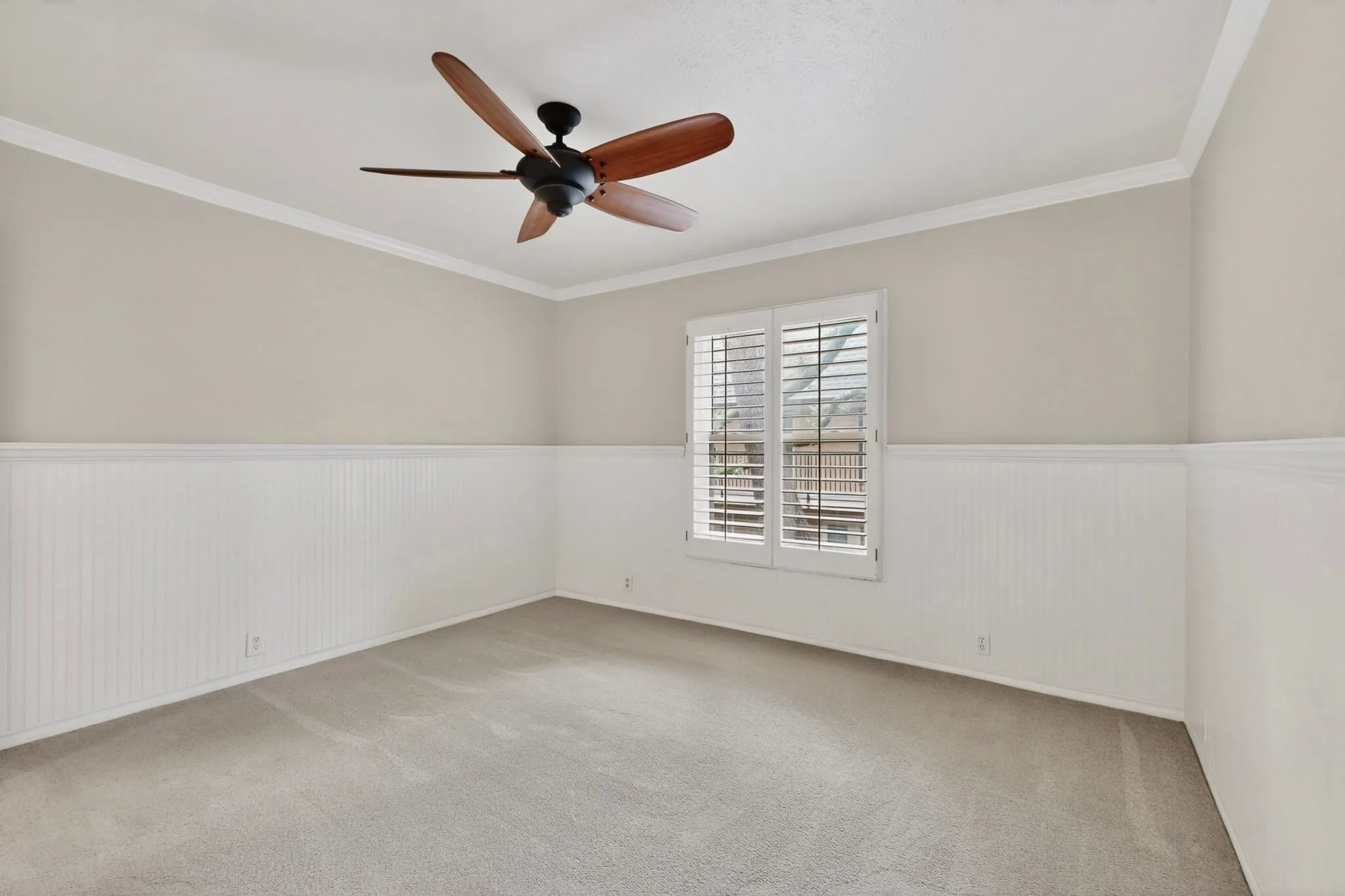 Carpeted interior room featuring a ceiling fan with wood-finish blades, crown molding, a window with plantation shutters, and beadboard wainscoting