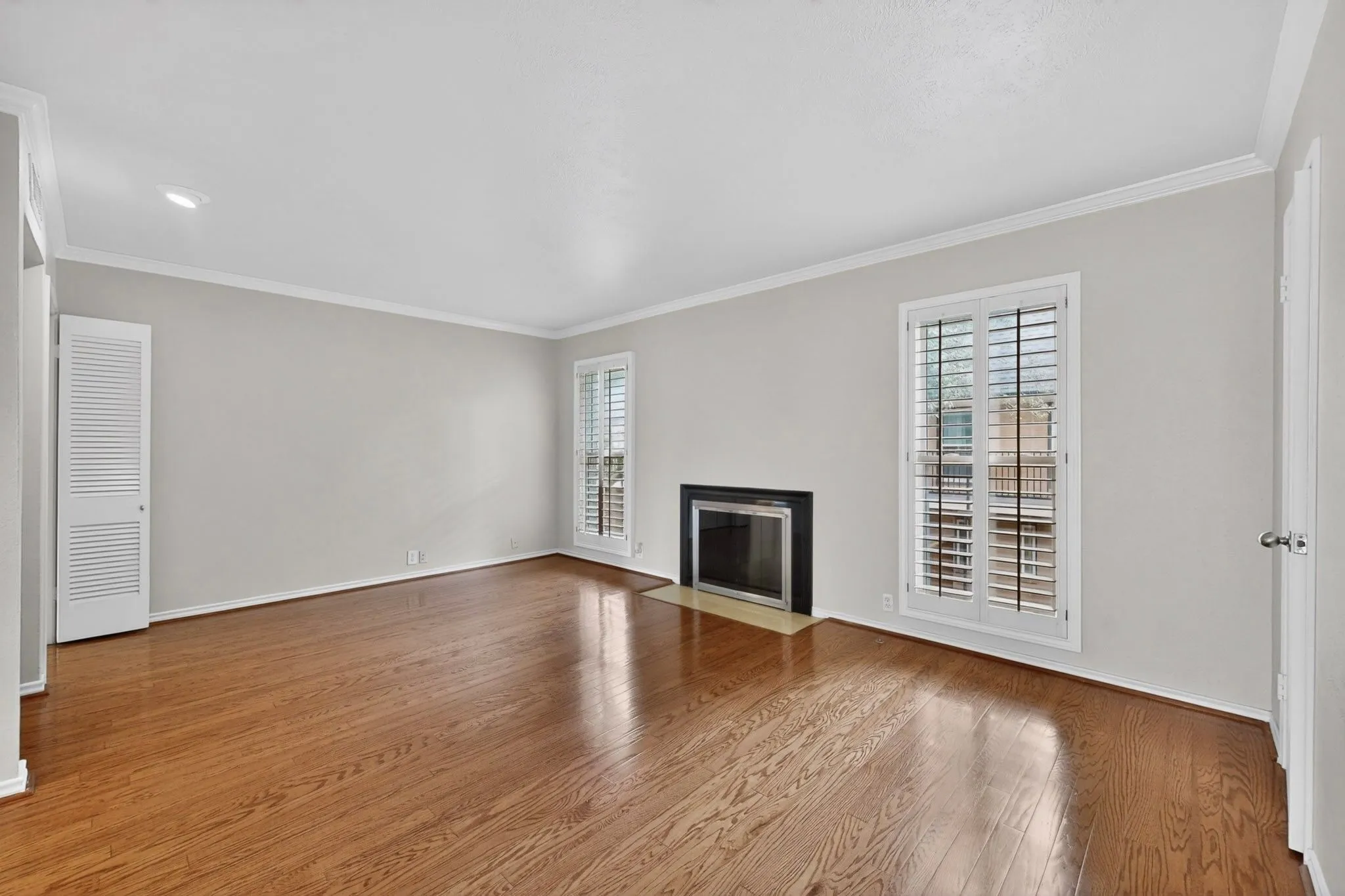 Spacious interior featuring wood-finish flooring, a recessed fireplace with a dark surround, dual windows with plantation shutters, crown molding, and a louvered bifold door