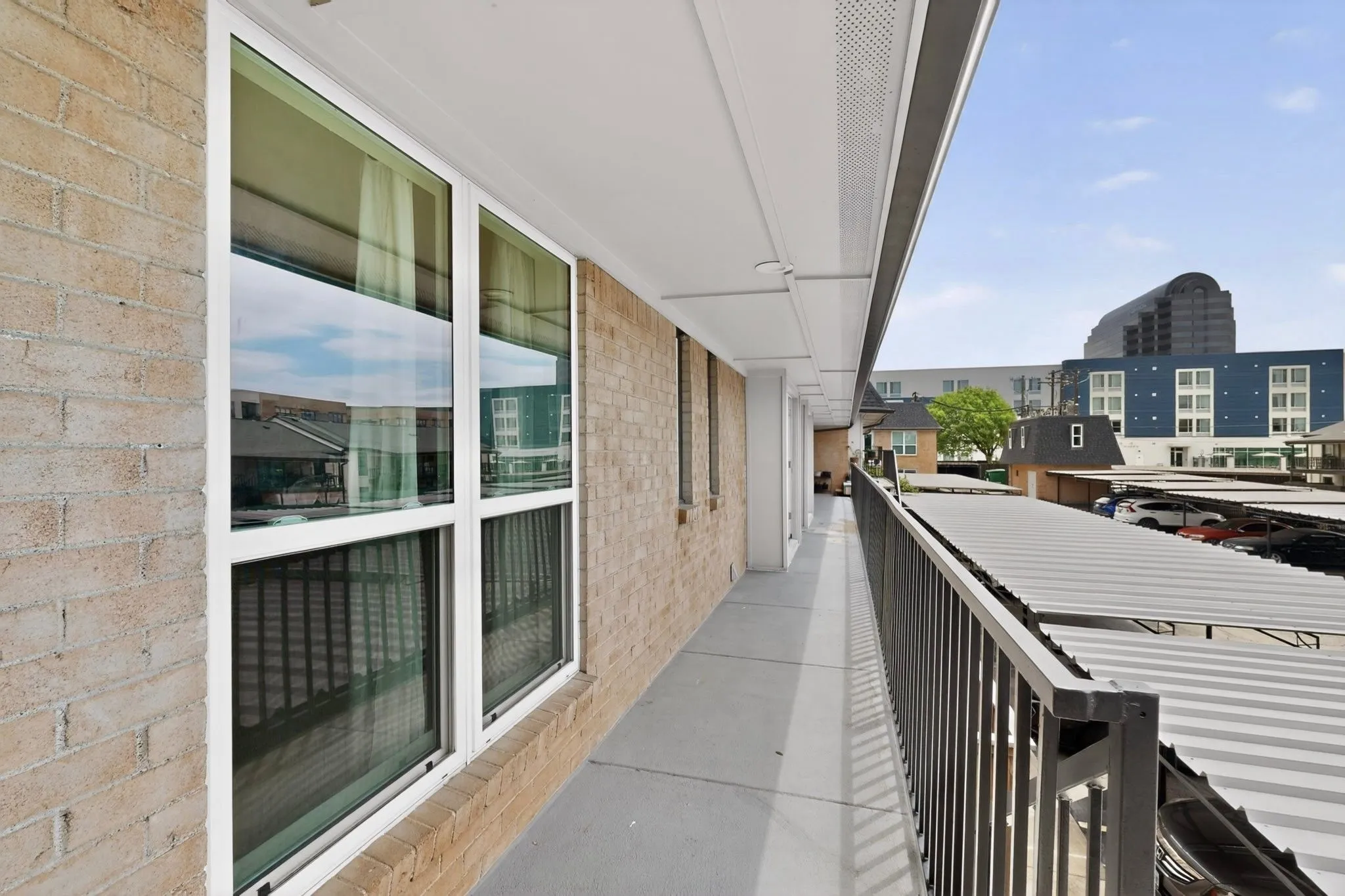Exterior walkway featuring a brick facade, white-framed windows, a concrete surface, and a black metal railing
