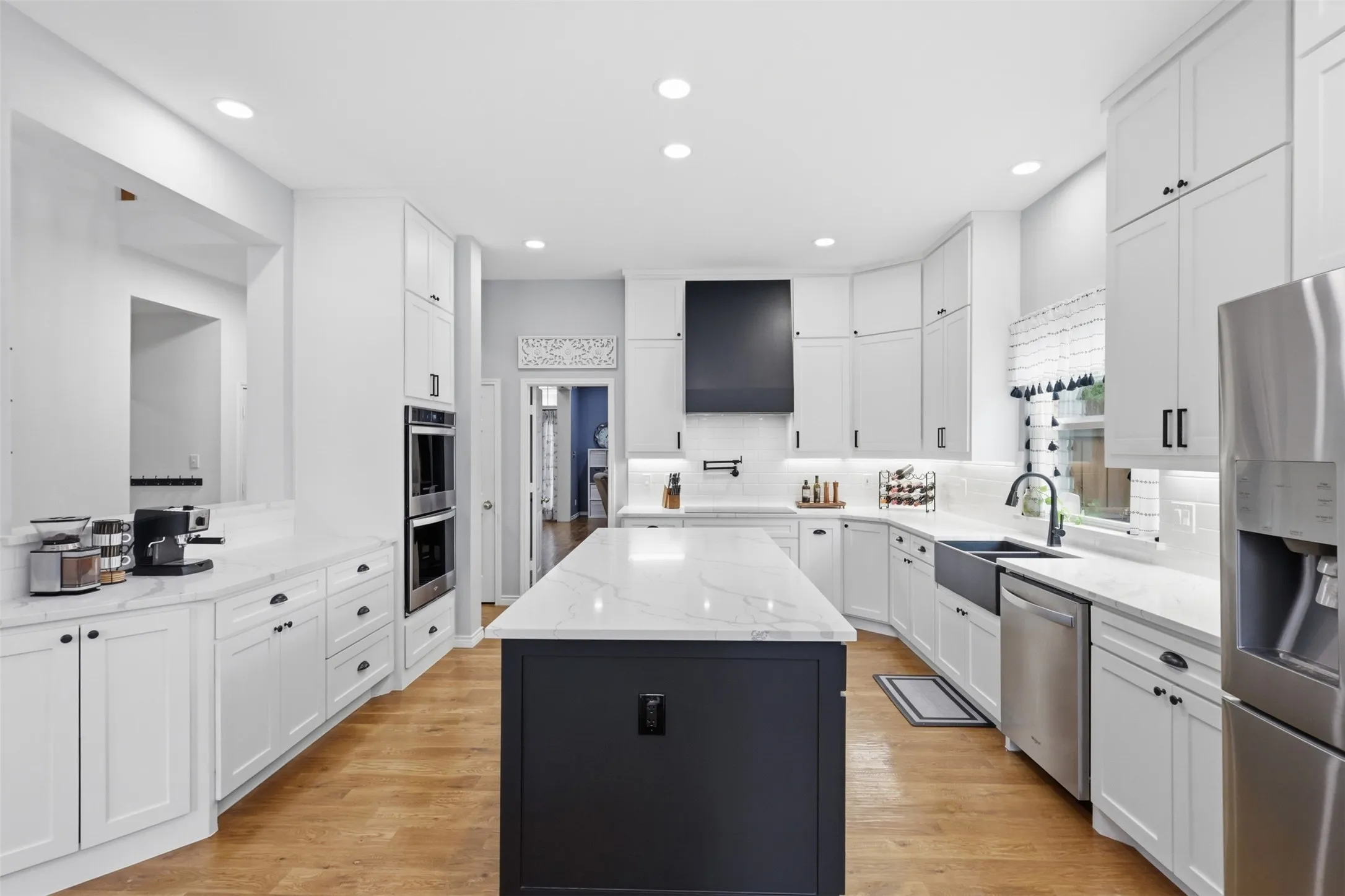 Spacious kitchen featuring a central island with a light-toned countertop, white cabinetry with dark hardware, recessed lighting, double wall ovens, and stainless steel appliances