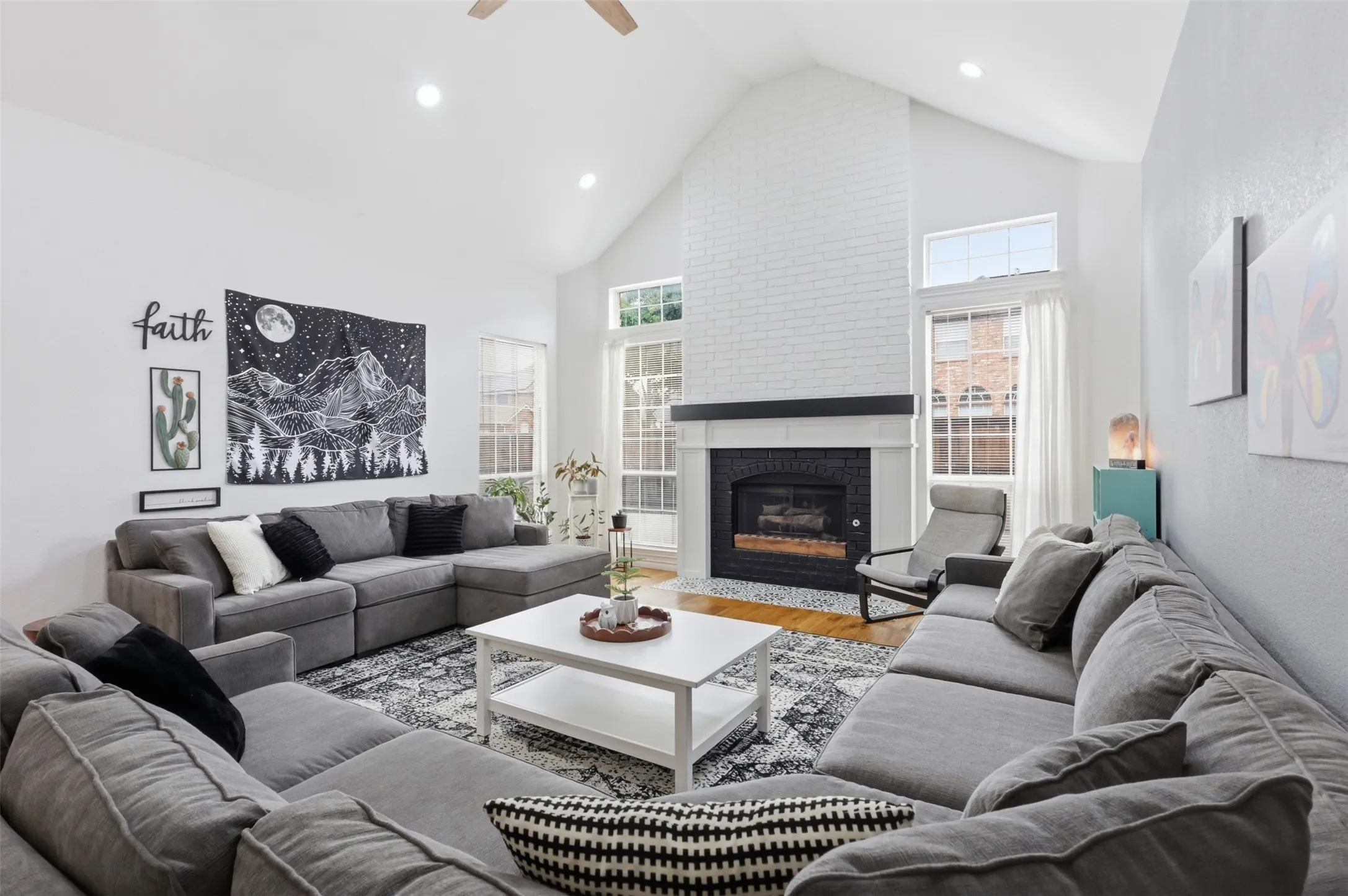 Living room featuring a vaulted ceiling with recessed lighting, a white brick fireplace with a dark mantel, multiple large windows, and wood-finish flooring
