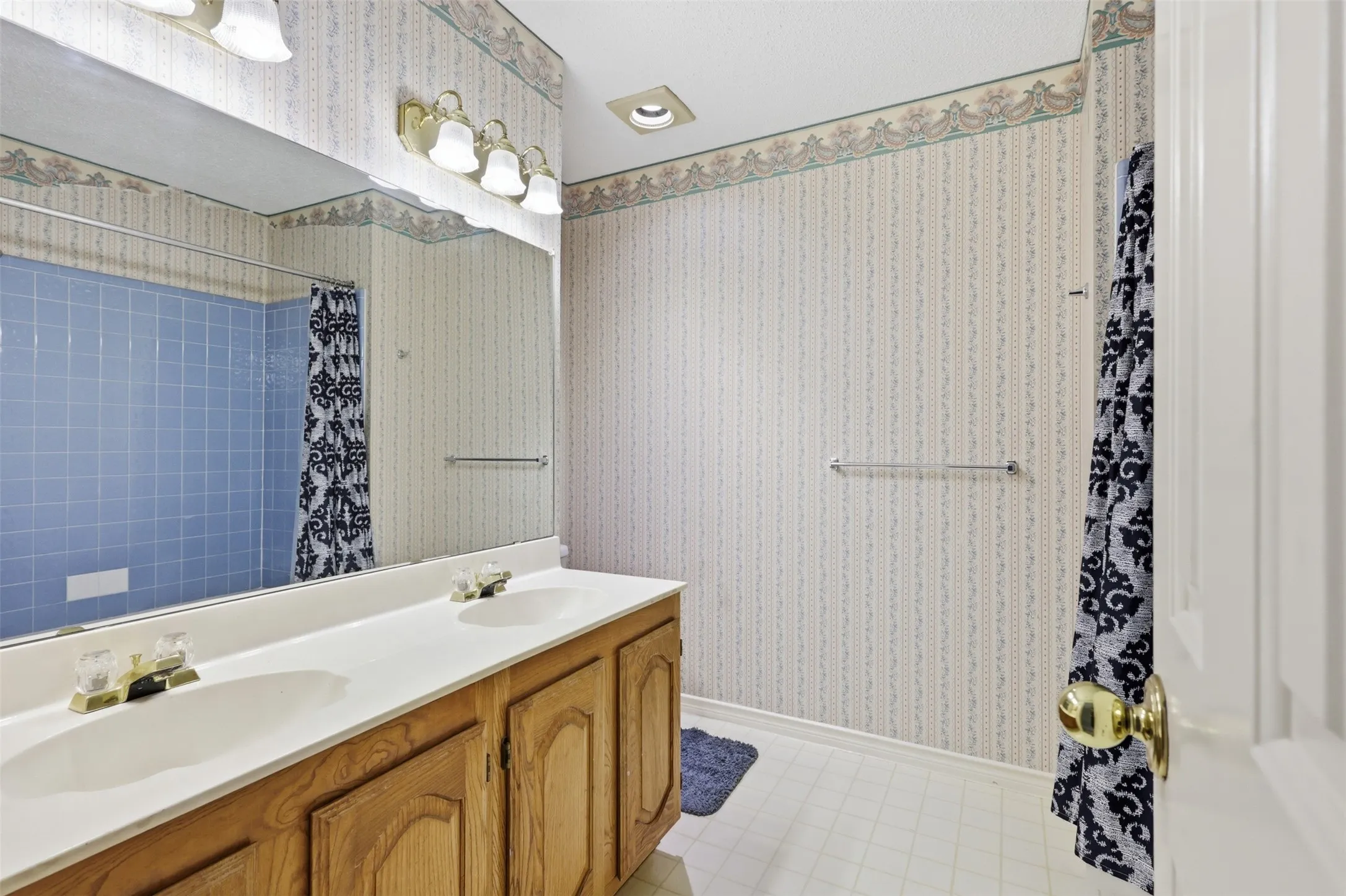 Bathroom featuring a double vanity with light wood cabinetry and integrated sinks, a large wall-mounted mirror, and a tiled shower enclosure