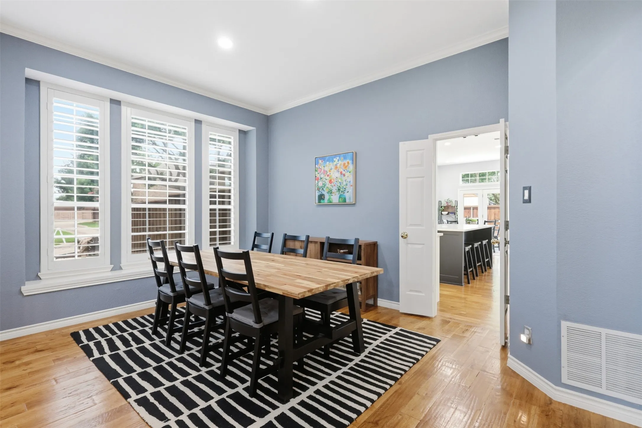 Dining area featuring light wood-finish flooring, three large windows with white louvered shutters, crown molding, and a light blue wall finish