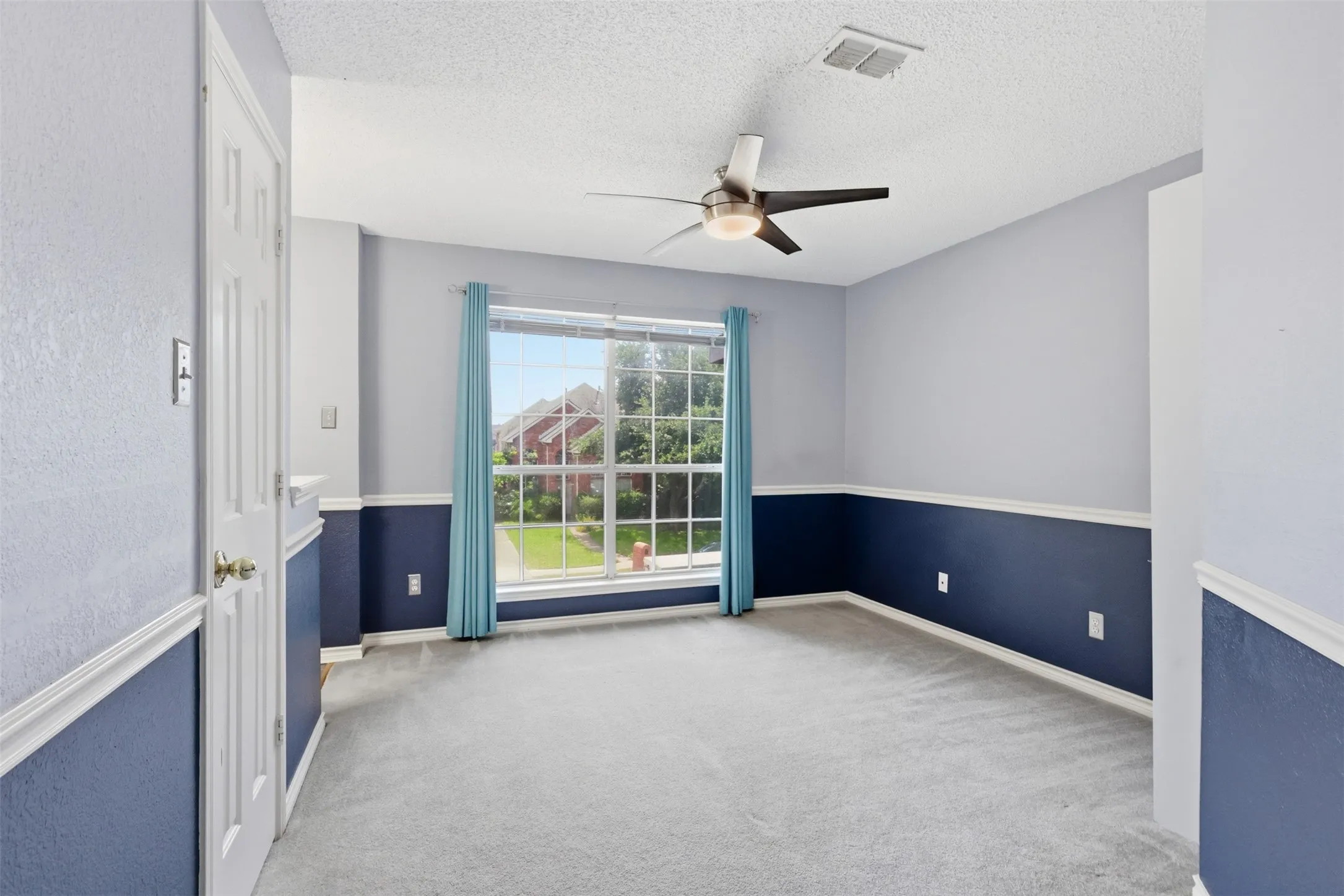 Spacious room featuring gray carpeting, a large multi-pane window with turquoise drapes, a ceiling fan with integrated lighting, and two-tone wall treatment with a chair rail