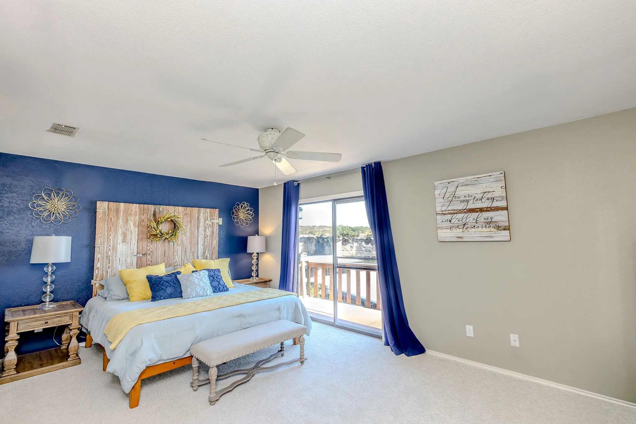 Carpeted room featuring a ceiling fan, a blue accent wall, and a sliding glass door providing outdoor access to a private balcony with wooden railings