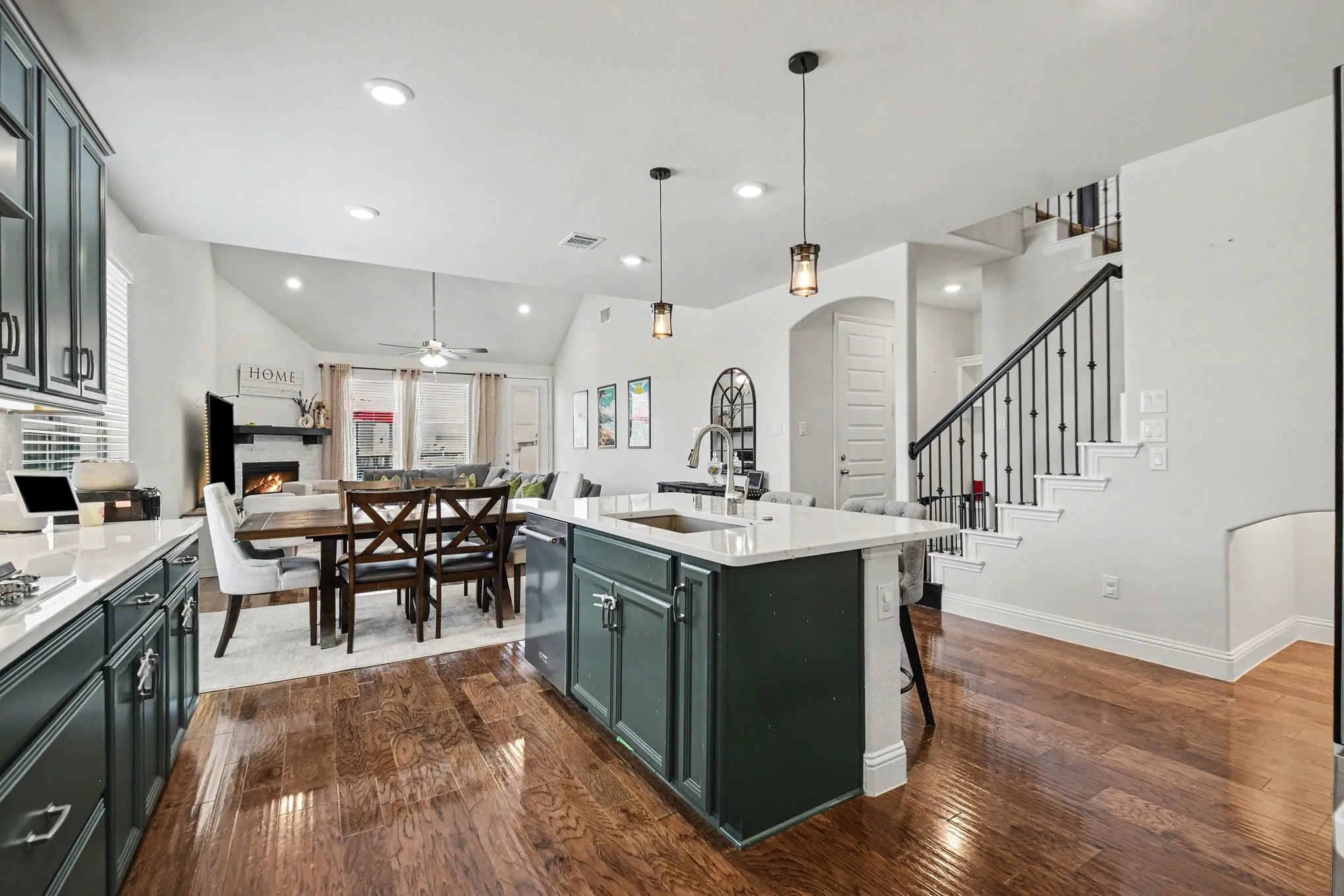 Open-concept living area featuring rich wood-finish flooring, a kitchen island with stone countertops, dark cabinetry, recessed lighting, and a prominent staircase with wrought iron balusters