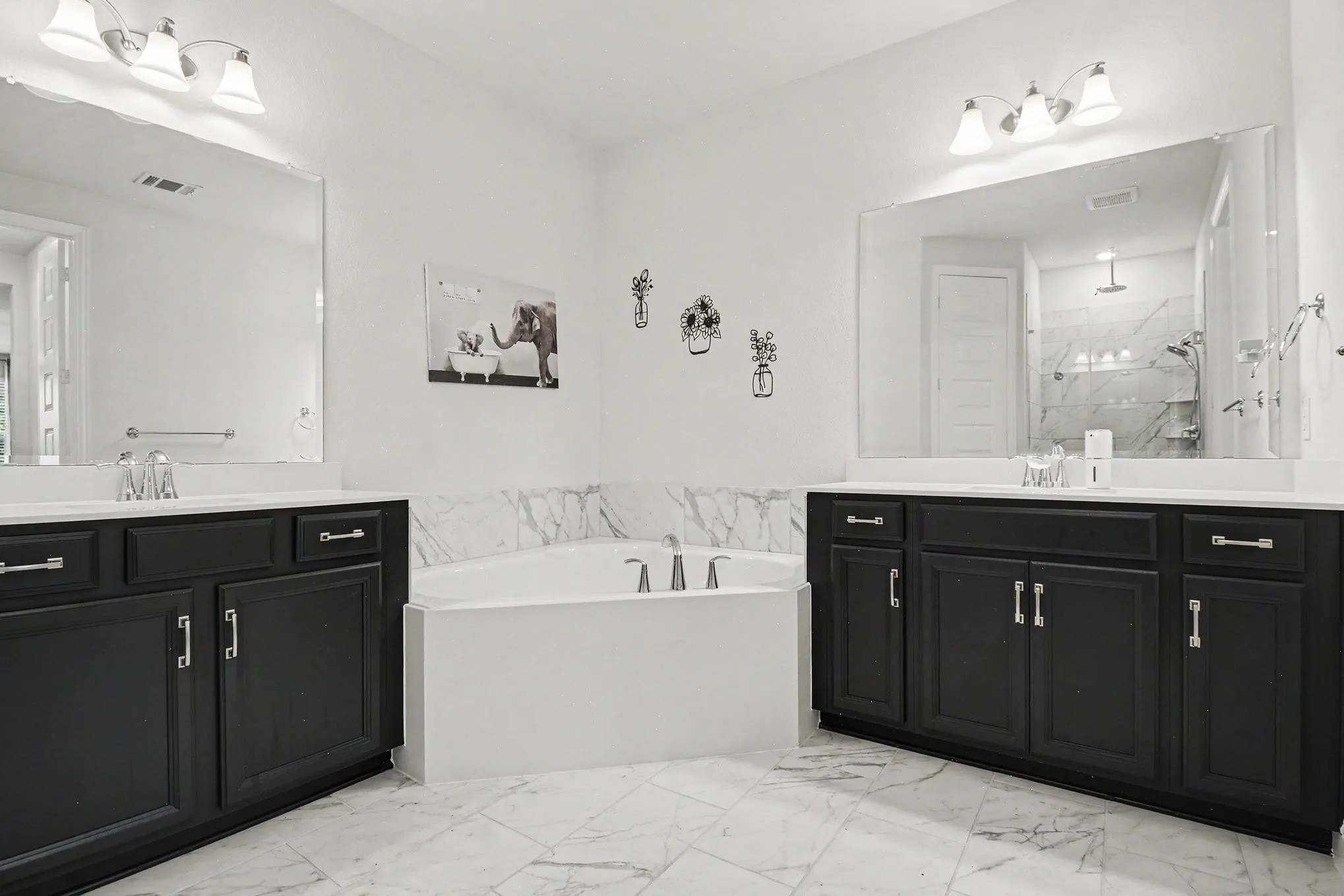 Soaking tub with surrounding marble-look tile, flanked by dual vanities with dark cabinetry and light countertops, under wall-mounted mirrors and sconces