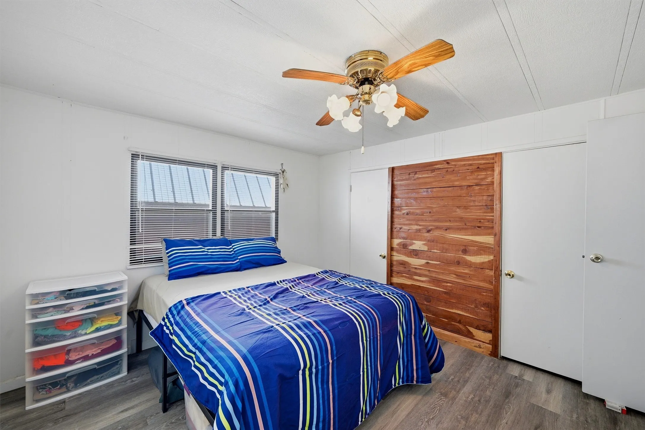 Room featuring wood-finish flooring, two windows with blinds, a ceiling fan with light fixture, and a built-in closet with sliding wooden doors