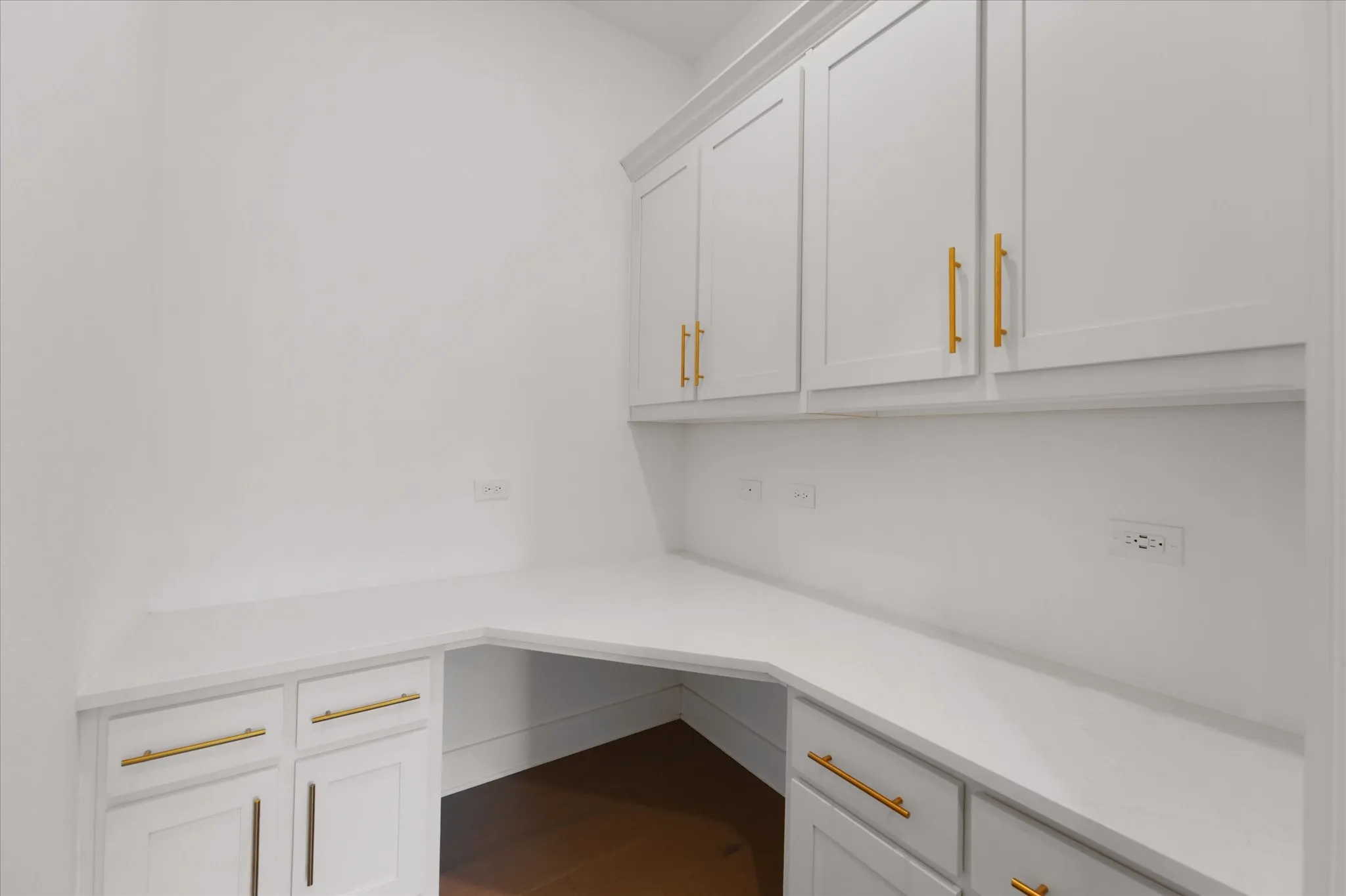 Built-in desk area featuring white cabinetry with gold-tone hardware, white countertops, upper storage cabinets, lower storage cabinets, and wood-finish flooring